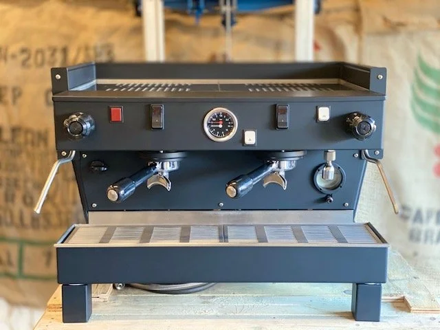 A black, commercial-grade coffee brewer with three spouts, control knobs, switches, and a gauge, placed on a wooden surface in a workshop or storage area.
