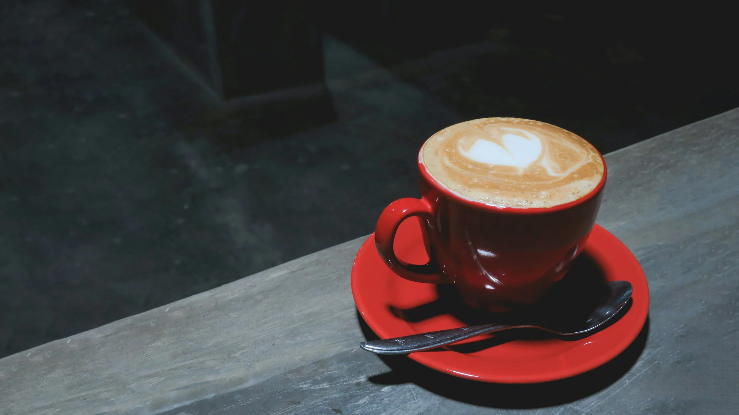 A red ceramic cup filled with latte art coffee on a matching red saucer with a silver spoon, placed on a gray wooden table.