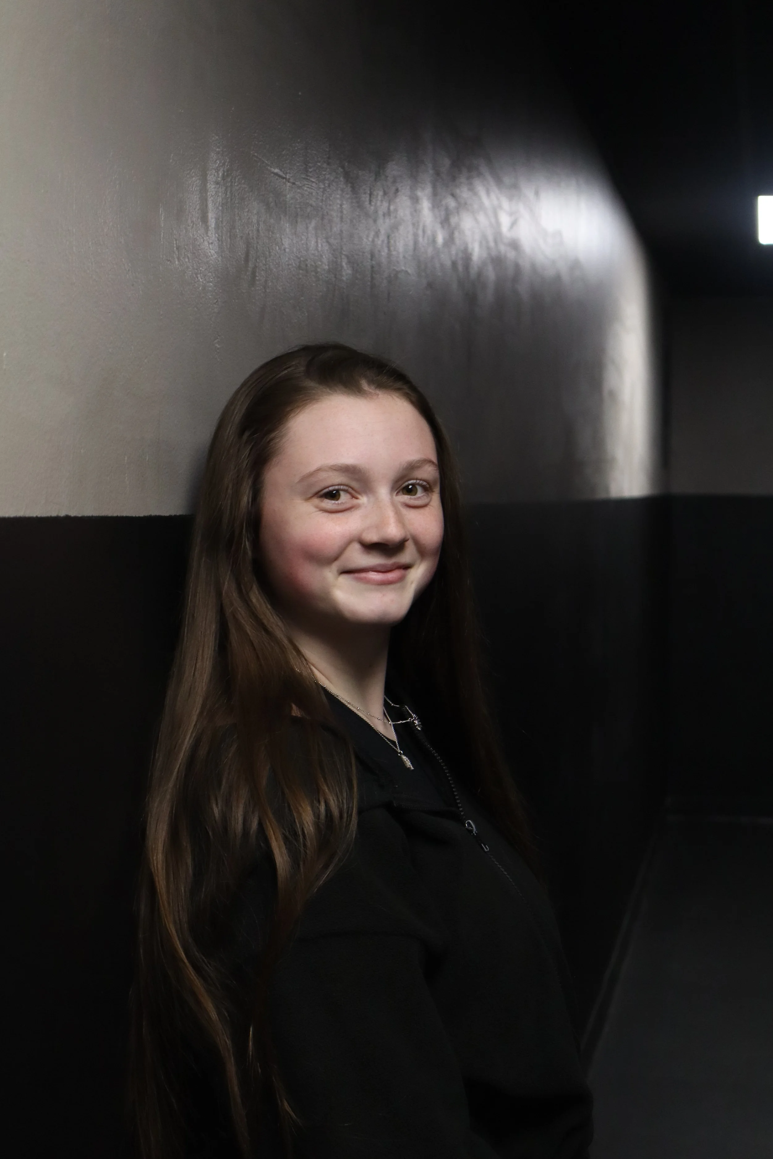 A young woman with long brown hair smiling while leaning against a dark wall in a dimly lit corridor.