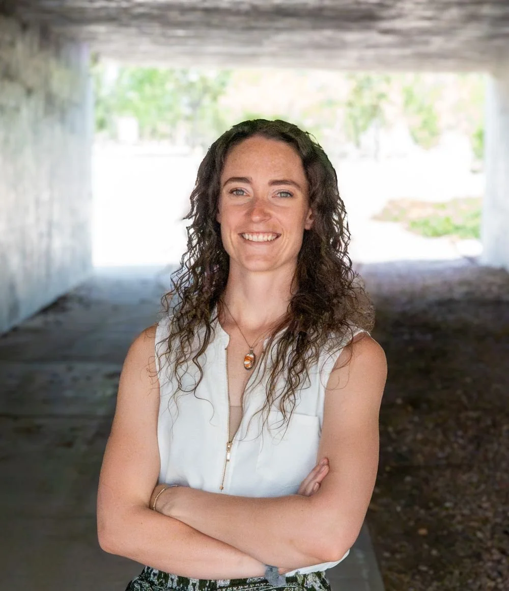 A woman with curly brown hair, wearing a sleeveless white top, stands with her arms crossed under a bridge, smiling at the camera.