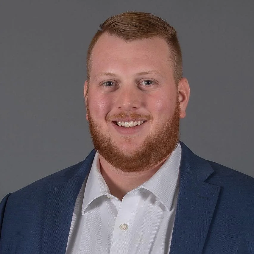 Headshot of a young man with short light brown hair and a beard, wearing a dark blue suit jacket and a white collared shirt, smiling against a gray background.