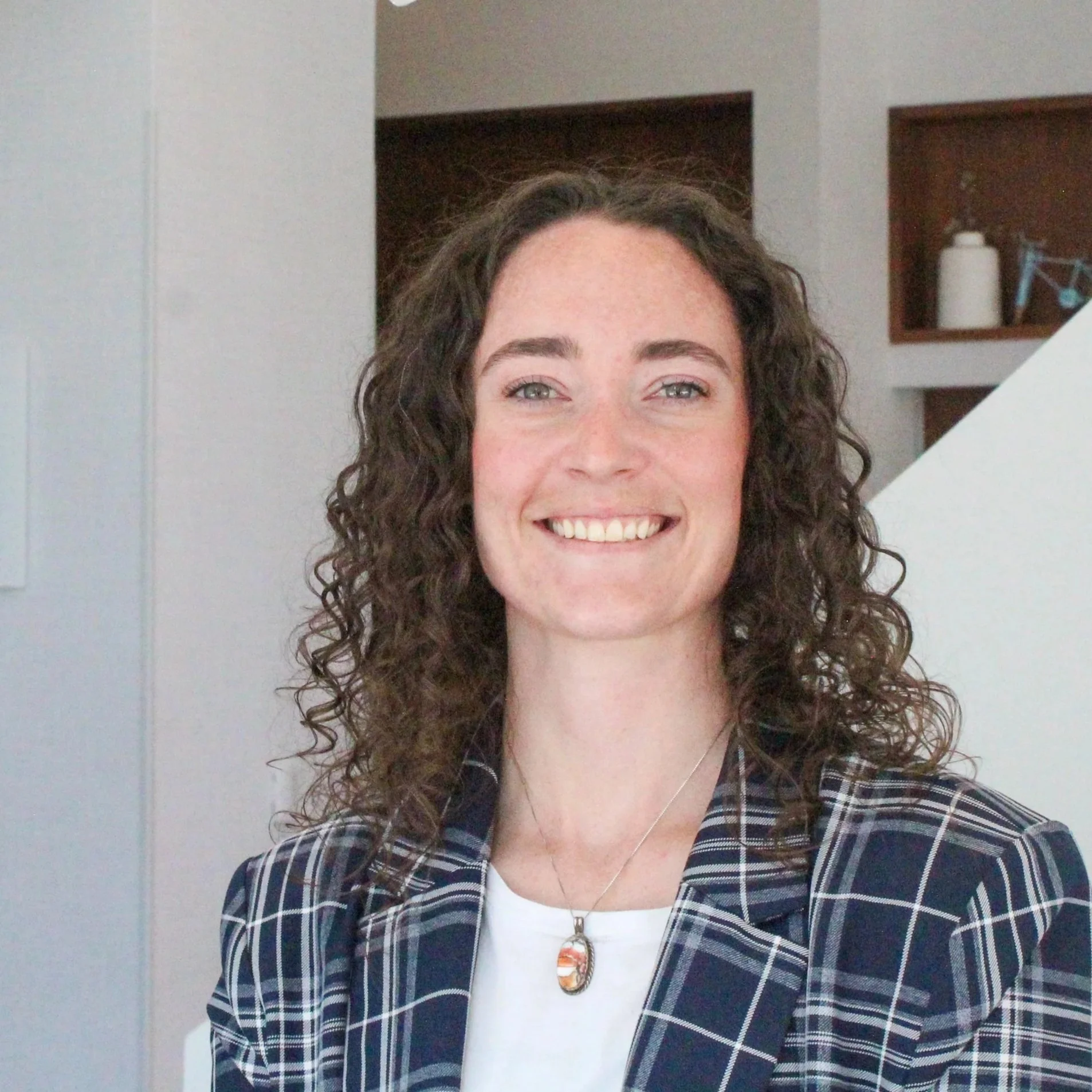 A smiling woman with curly brown hair, wearing a plaid blazer and a necklace, standing indoors in front of a modern background with shelves.