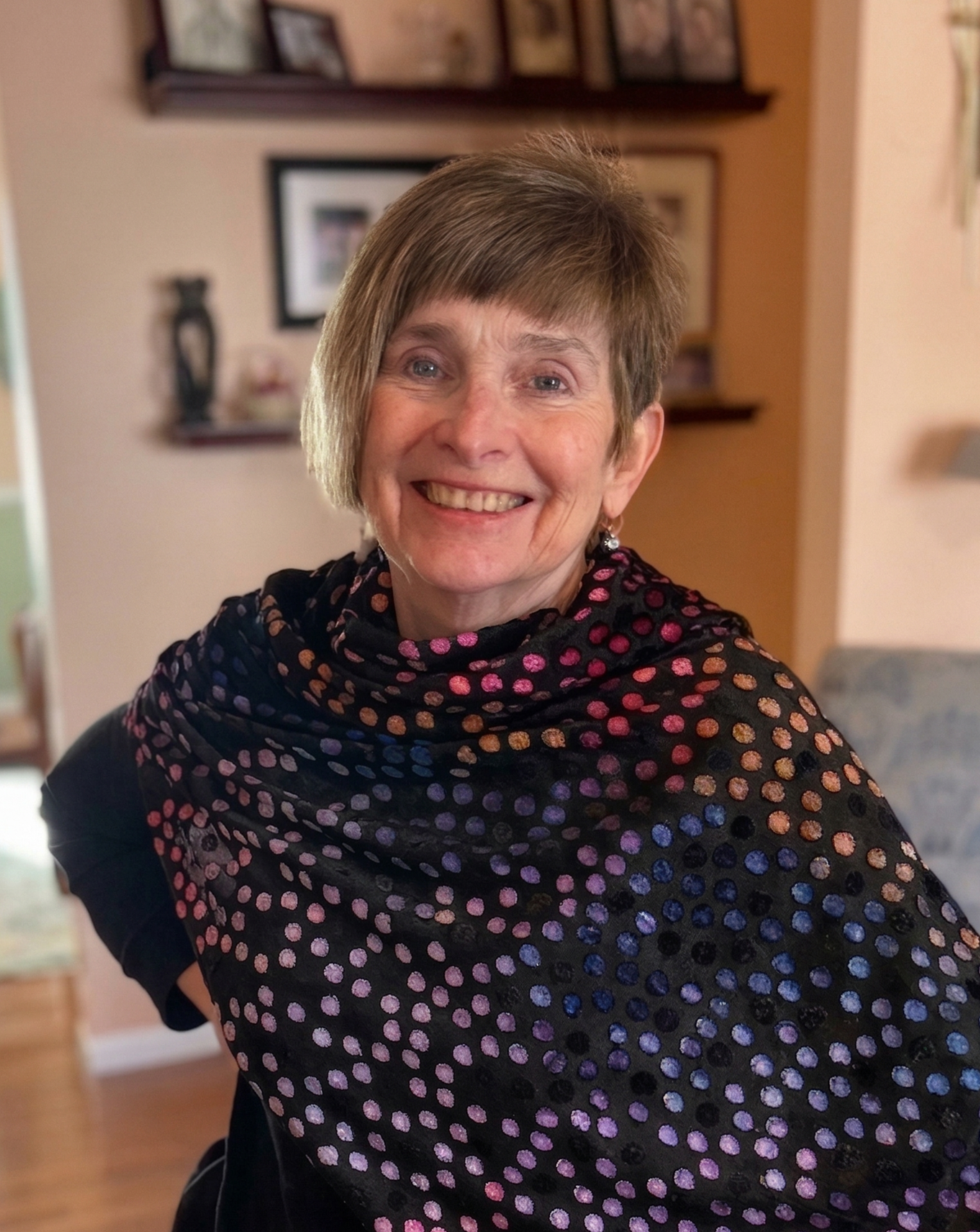 A smiling woman with short hair, wearing a black shawl with colorful polka dots, standing inside a home with shelves and framed pictures in the background.