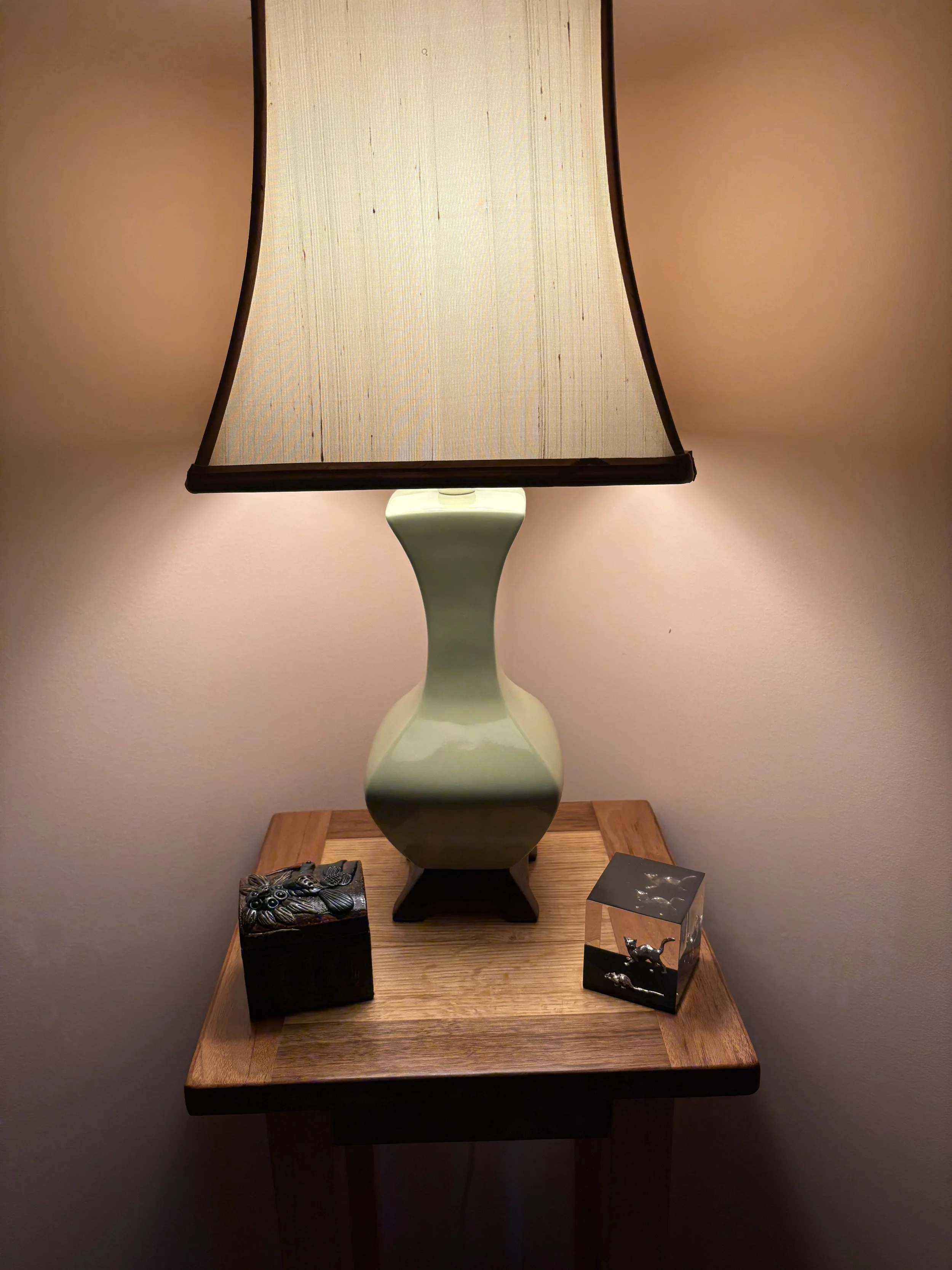 A table lamp with a green ceramic base and a cream lampshade sits on a wooden side table, with two small decorative boxes nearby.