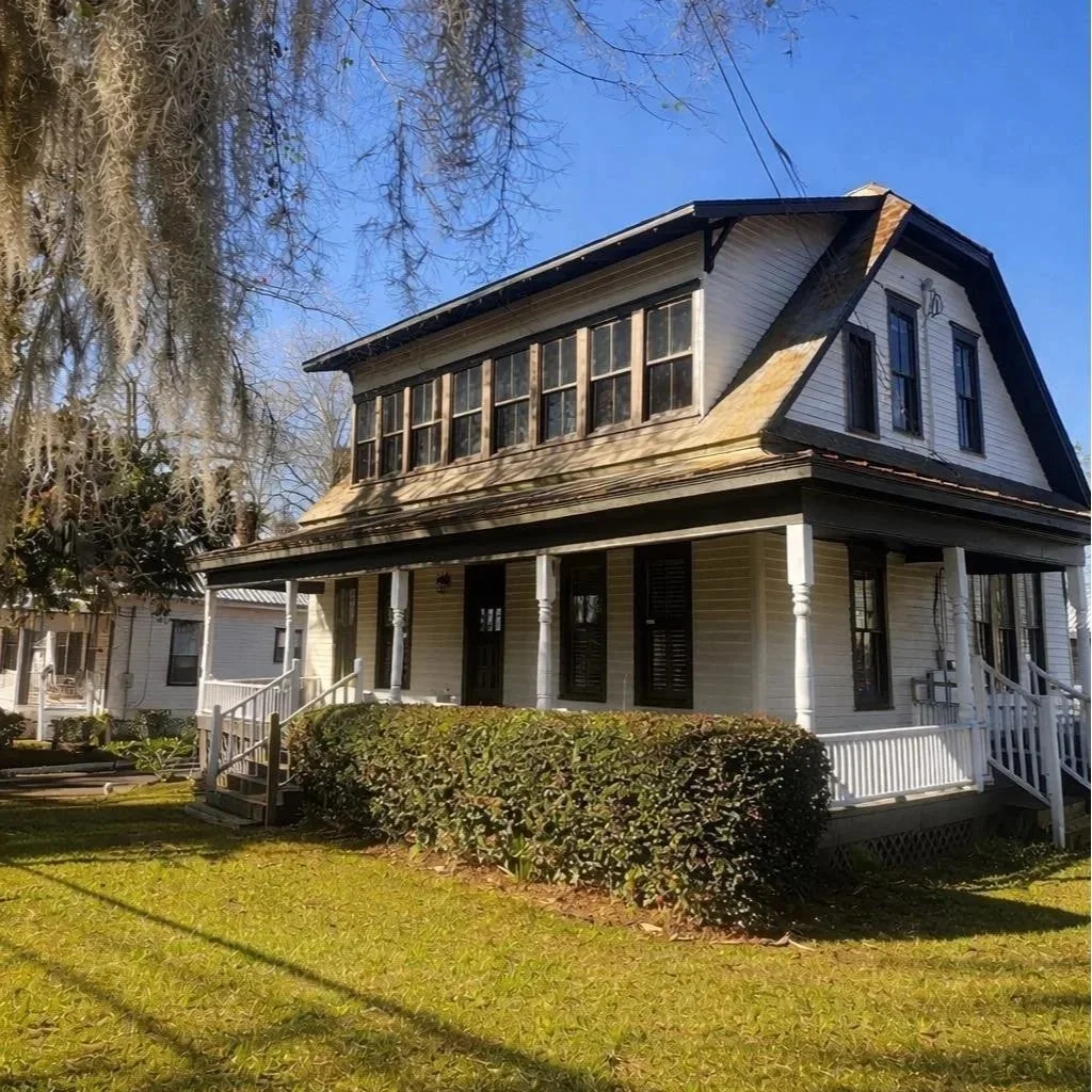 A two-story house with a covered front porch, white siding, black shutters, and a steep gabled roof. The house features enclosed second-story windows and a well-maintained lawn with bushes in front.