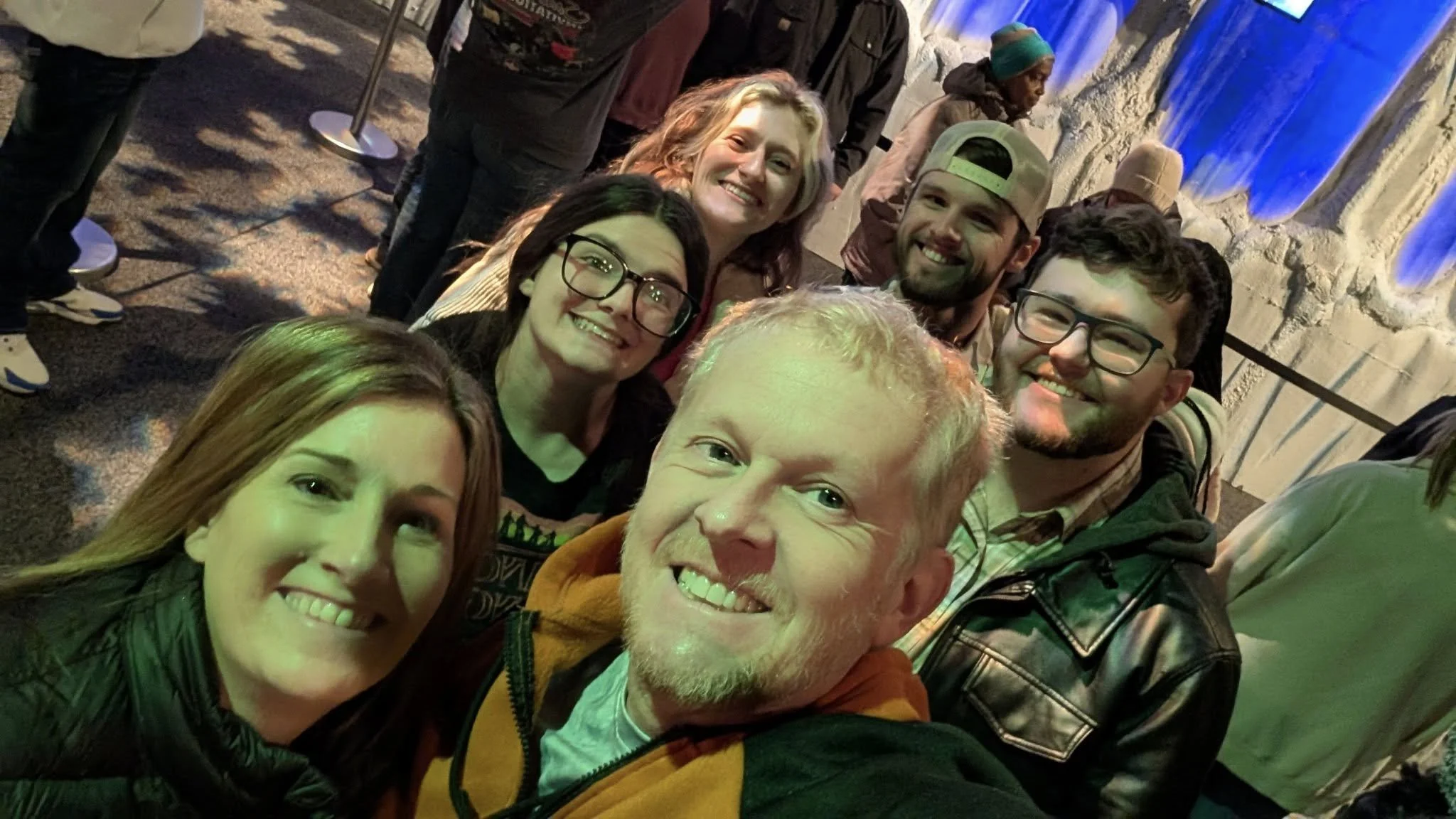 Group of six smiling young adults taking a selfie indoors, with artificial lighting and colorful wall decorations in the background.
