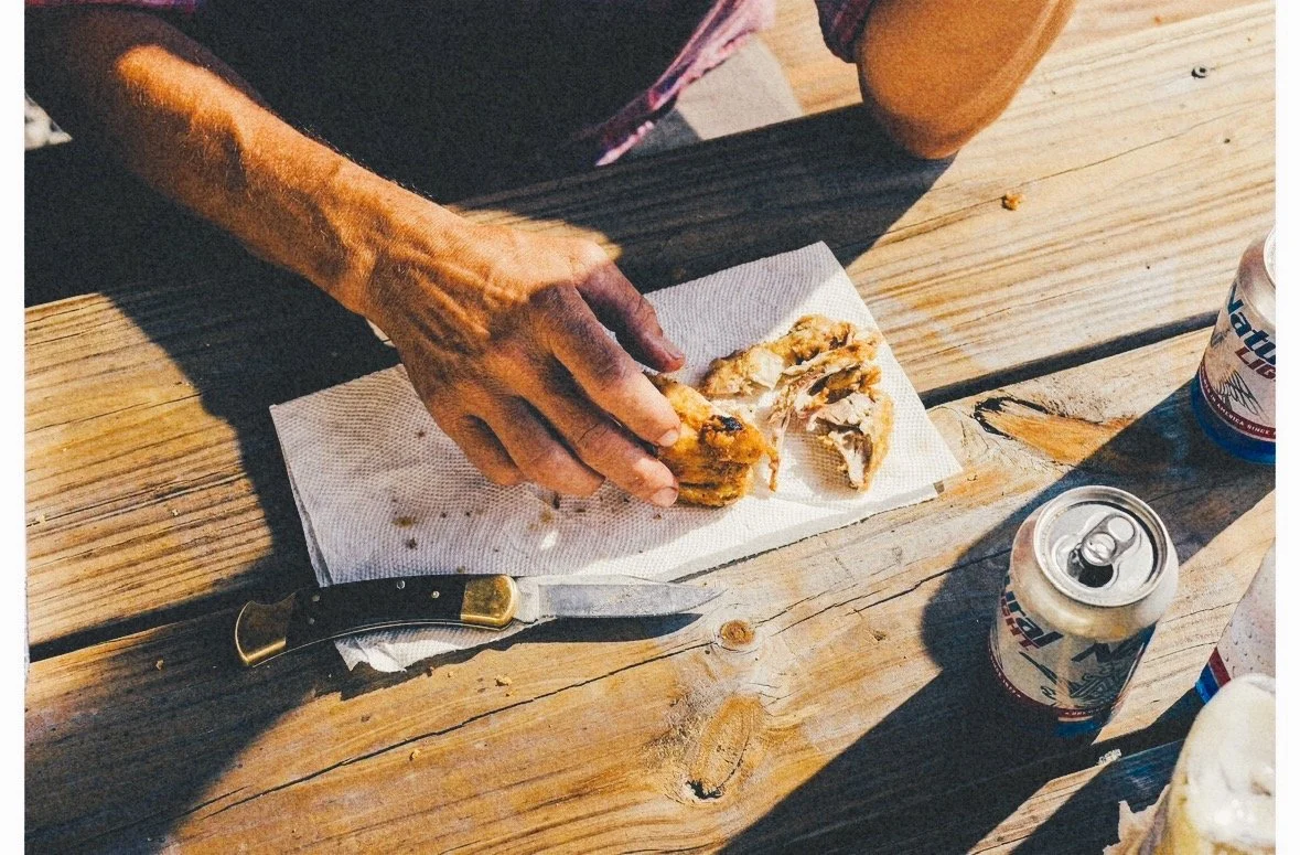 A person tearing apart fried chicken on a napkin on a wooden table, with a folding knife and two cans of beer nearby.