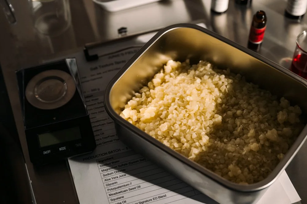 Crushed beeswax or paraffin wax in a metal container, on a kitchen countertop with a small digital scale, a printed recipe, and small bottles of essential oils in the background.