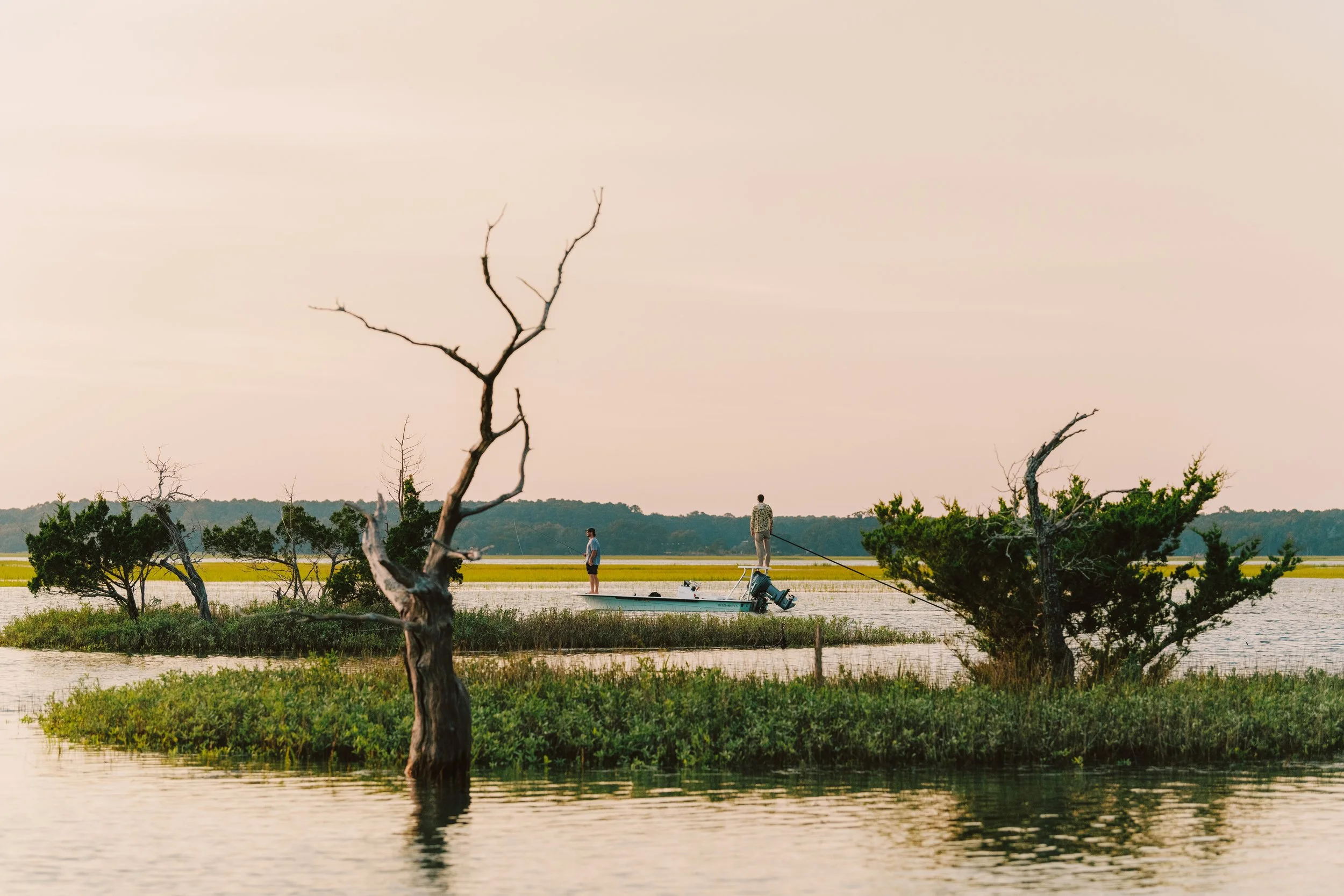 Two people fishing on a boat in a marshy area with trees, water, and fields in the background during sunset.