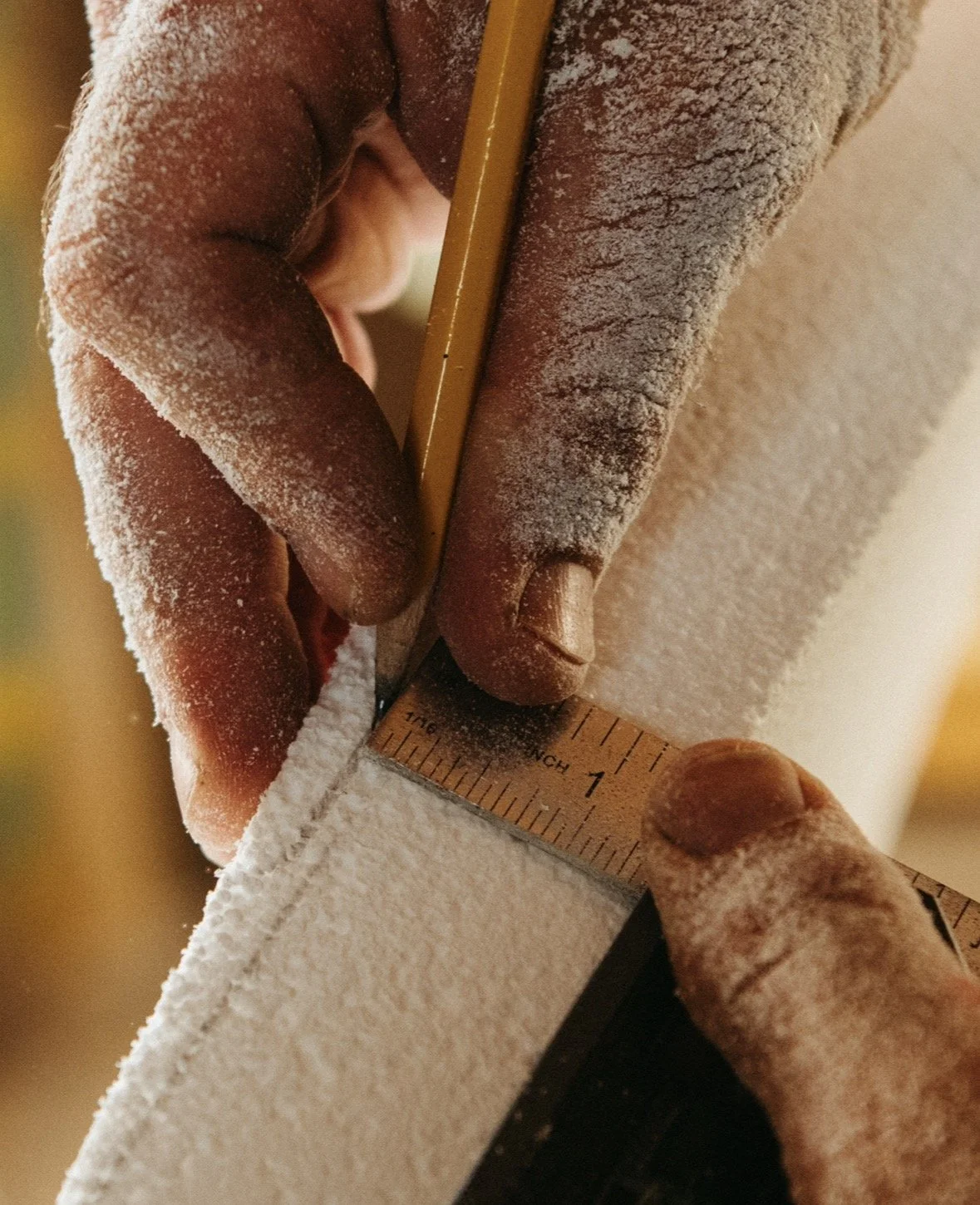 Close-up of a person's hand holding a ruler and a pencil, measuring and marking a piece of white foam or insulation.