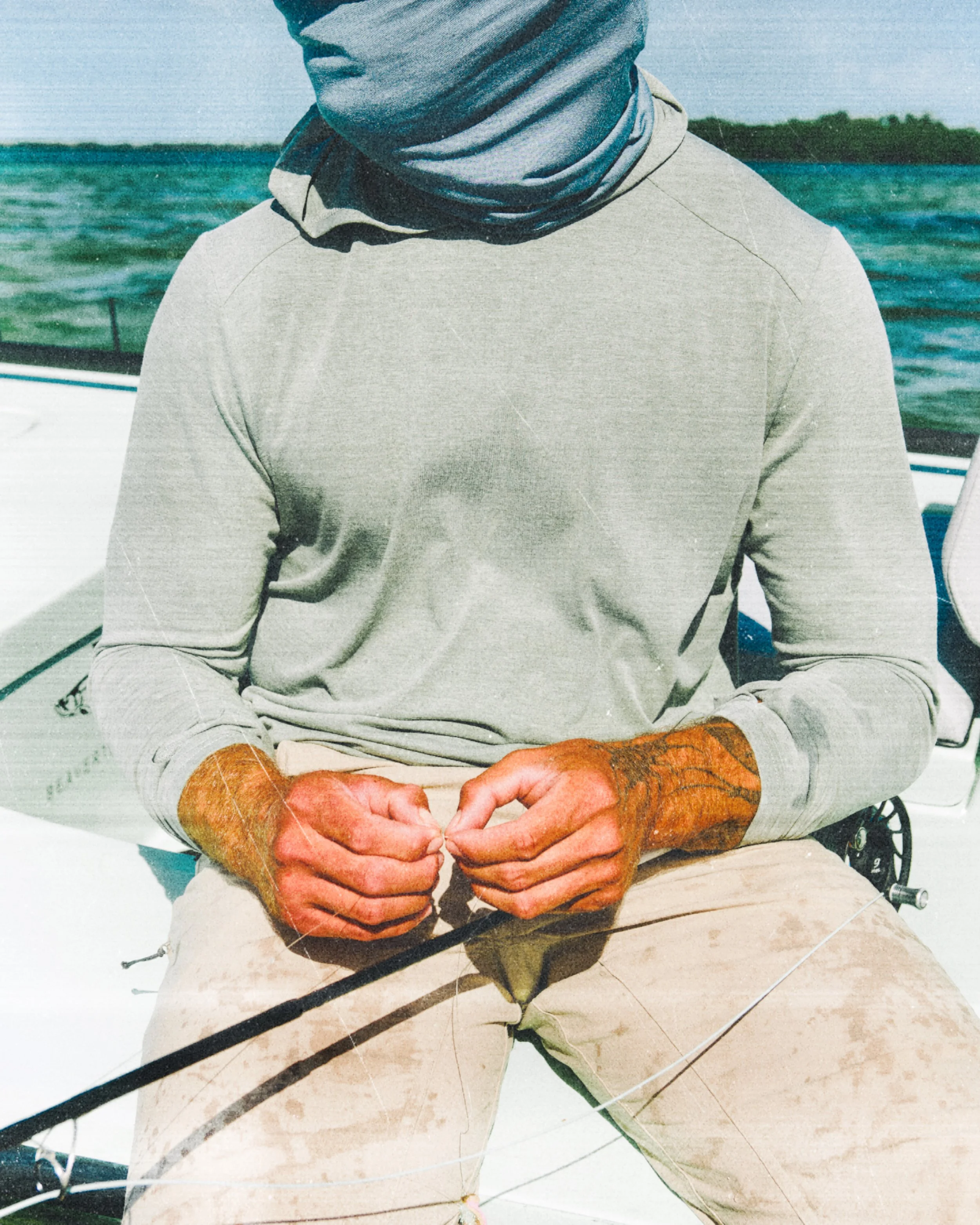Person sitting on a boat, wearing a long-sleeve shirt and light-colored pants, with hands operating fishing line, near water and shoreline.