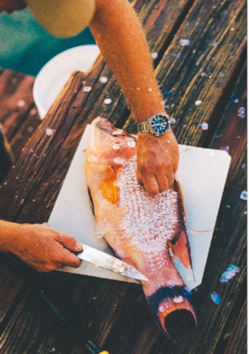 Person preparing to fillet a fish on a cutting board, with a knife in hand, on a wooden deck with scattered confetti.