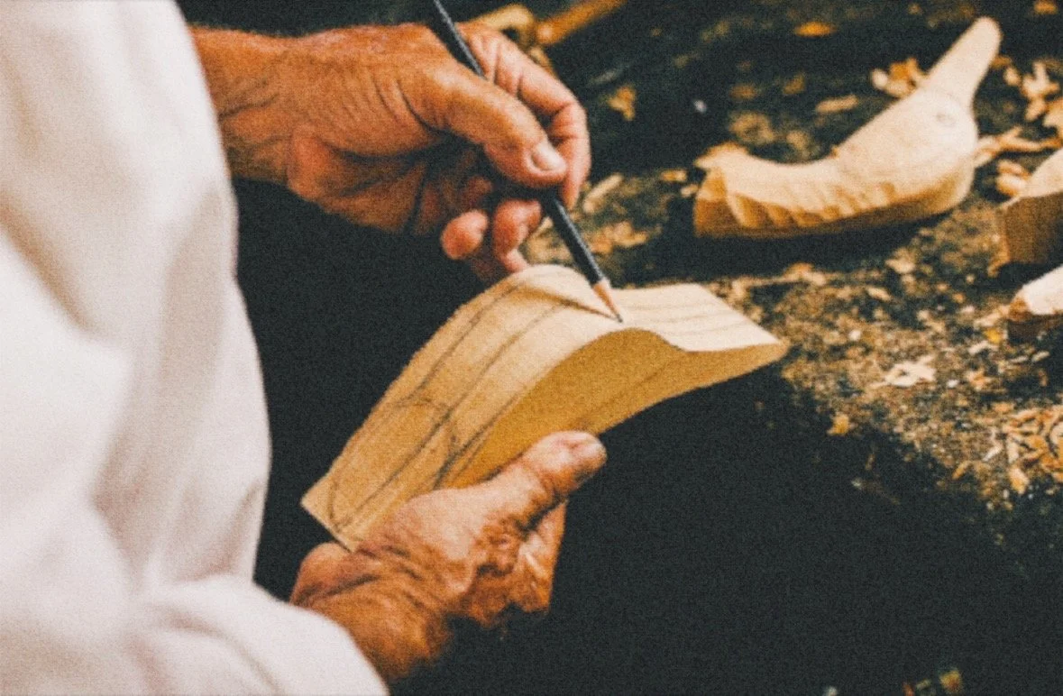 A person carving wood with a pencil, using their hand to steady the piece of wood in a woodworking shop.