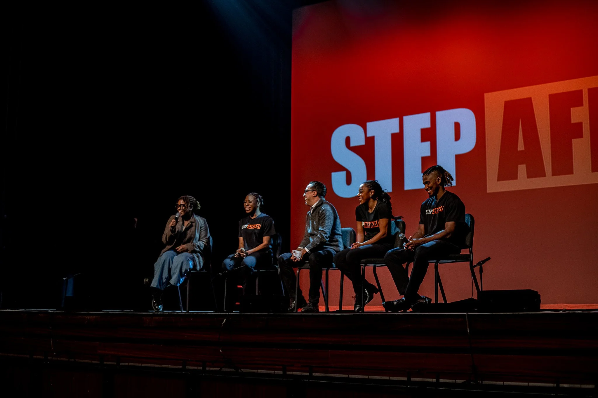 Five individuals sitting on chairs on stage during a panel discussion, with a large screen behind them displaying the word 'STEP' and part of a sign that says 'AFRIKA!'