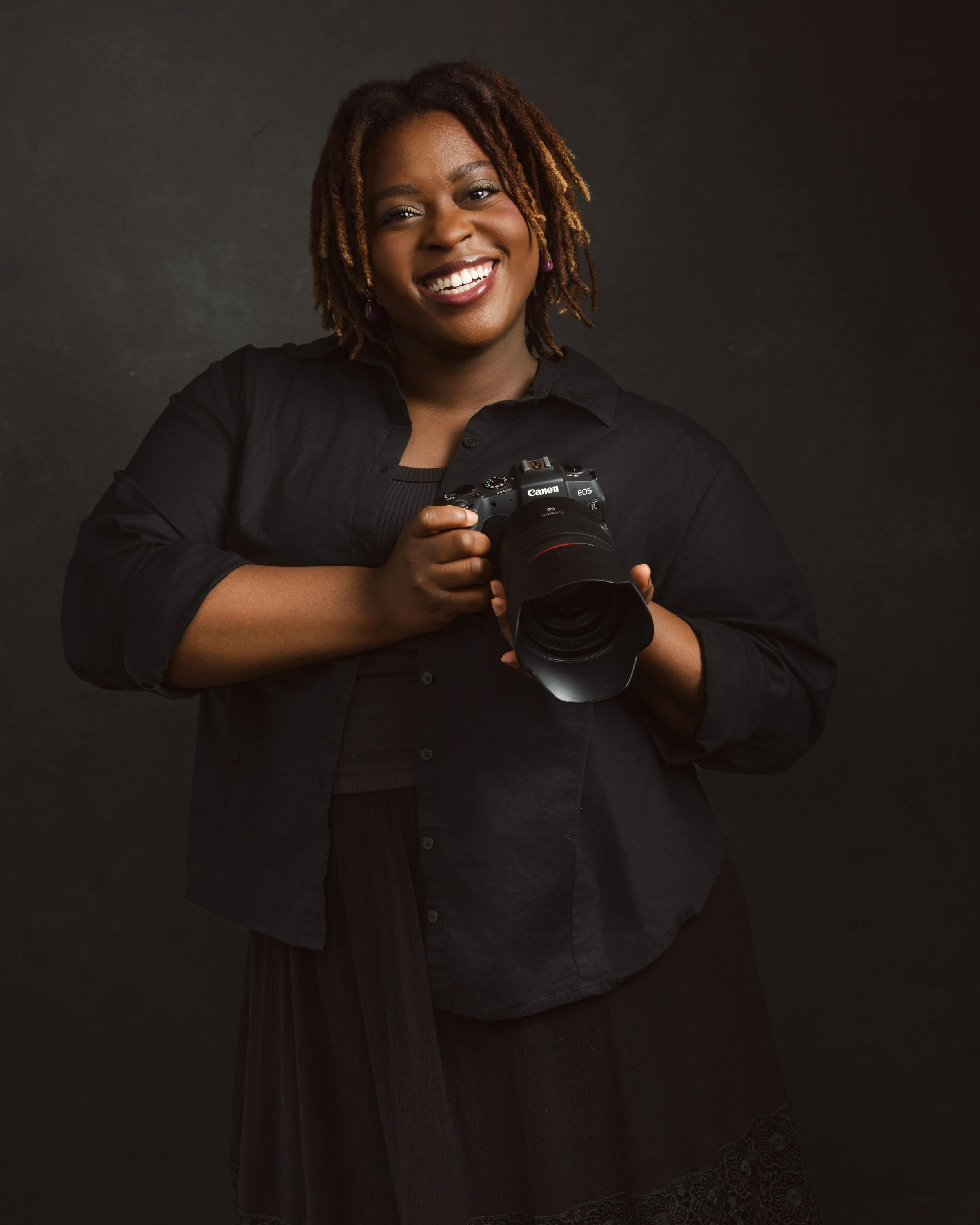 A woman with dreadlocks holding a Canon camera and smiling.