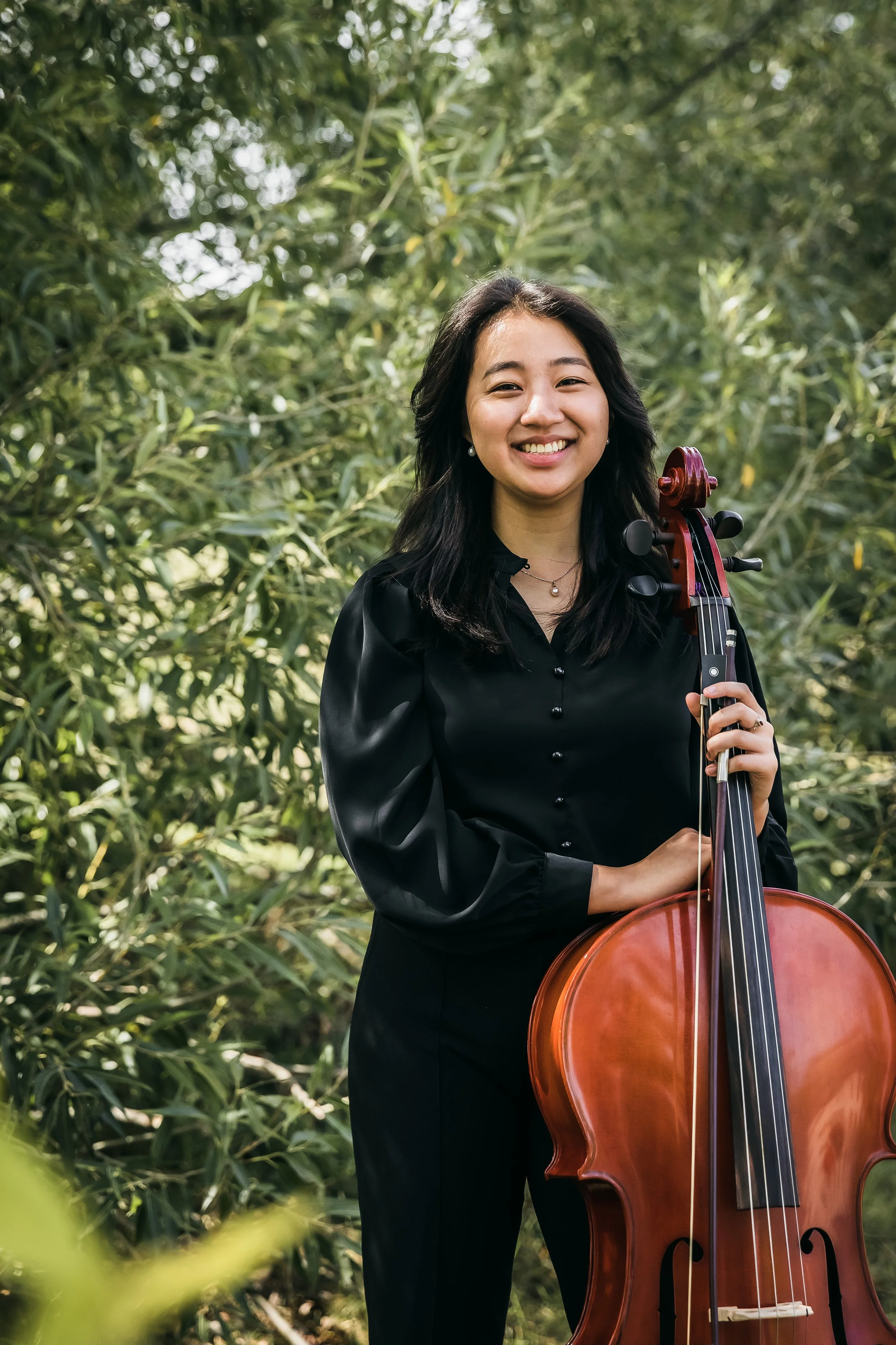 A woman in black clothing holding a cello in an outdoor setting with green trees and bushes.