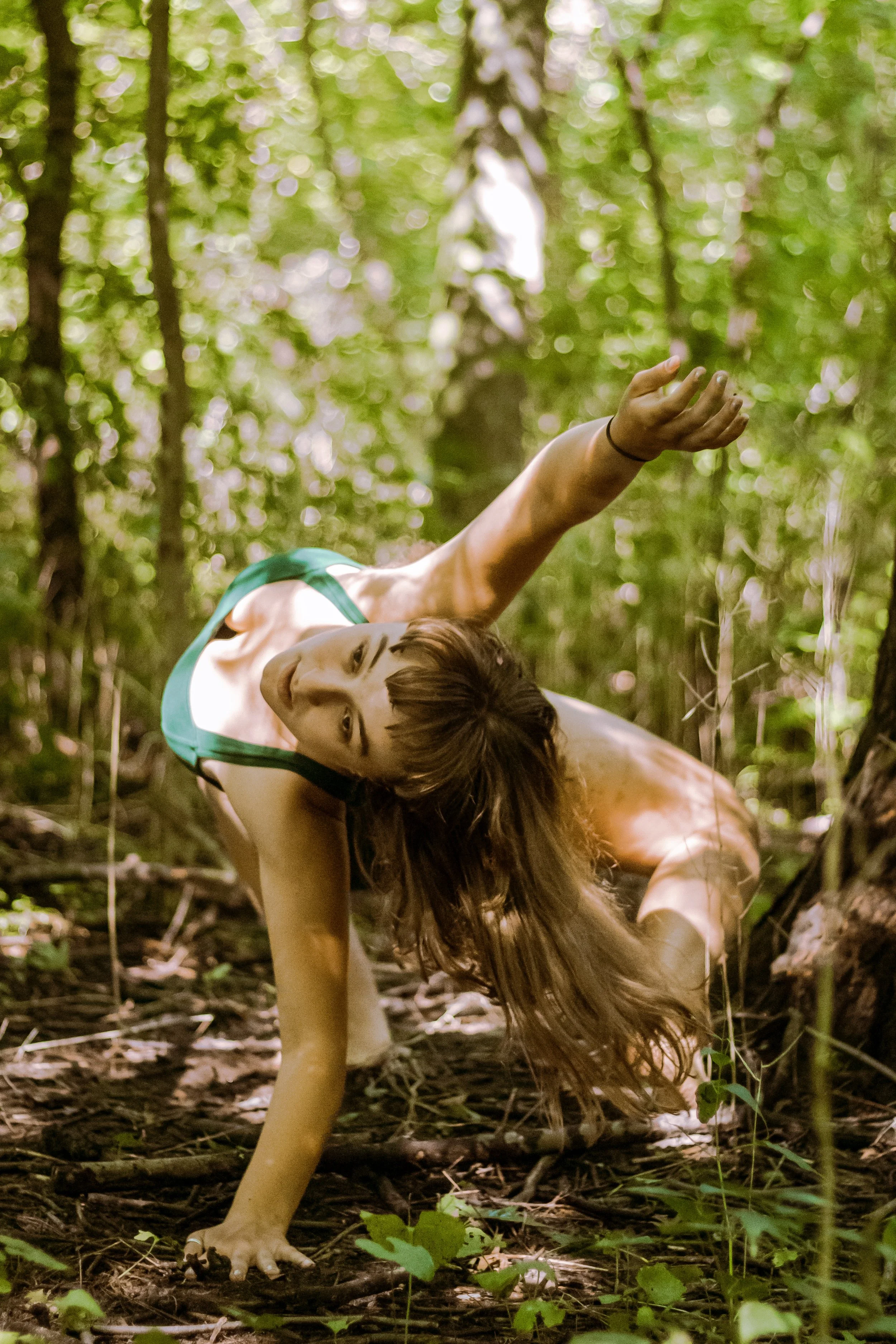 A woman performing a yoga pose outdoors in a lush green forest, balancing on one hand with her hair flowing downward and her other arm extended upward.
