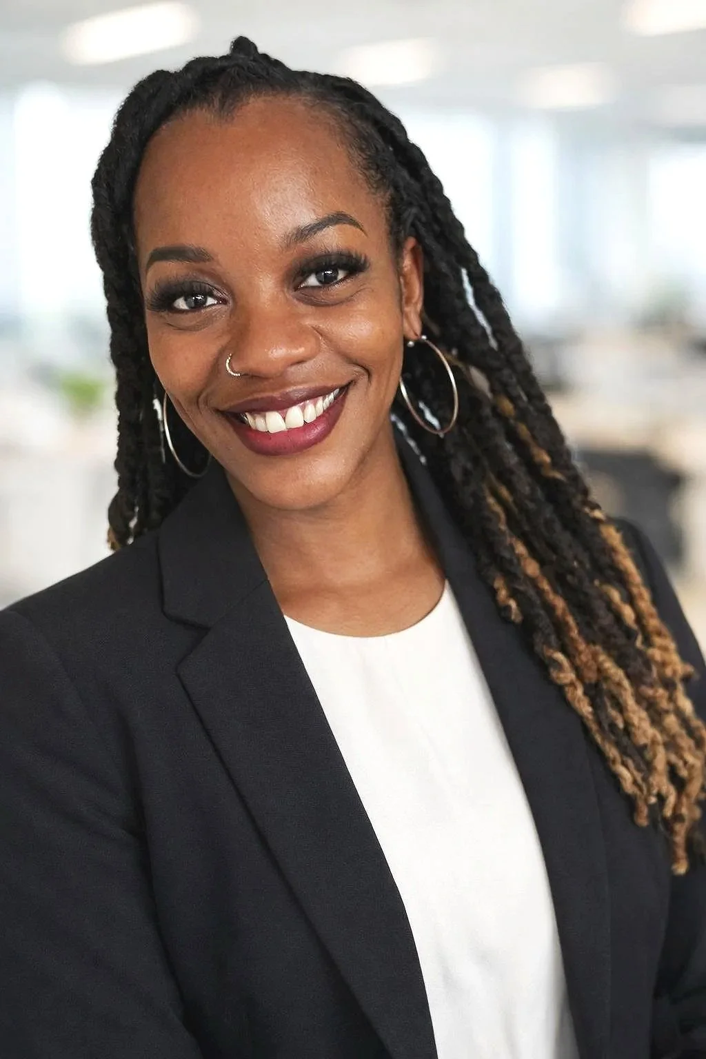 A confident woman with dark skin, long textured hair styled in twists, smiling, wearing a black blazer, white top, hoop earrings, in an office setting.
