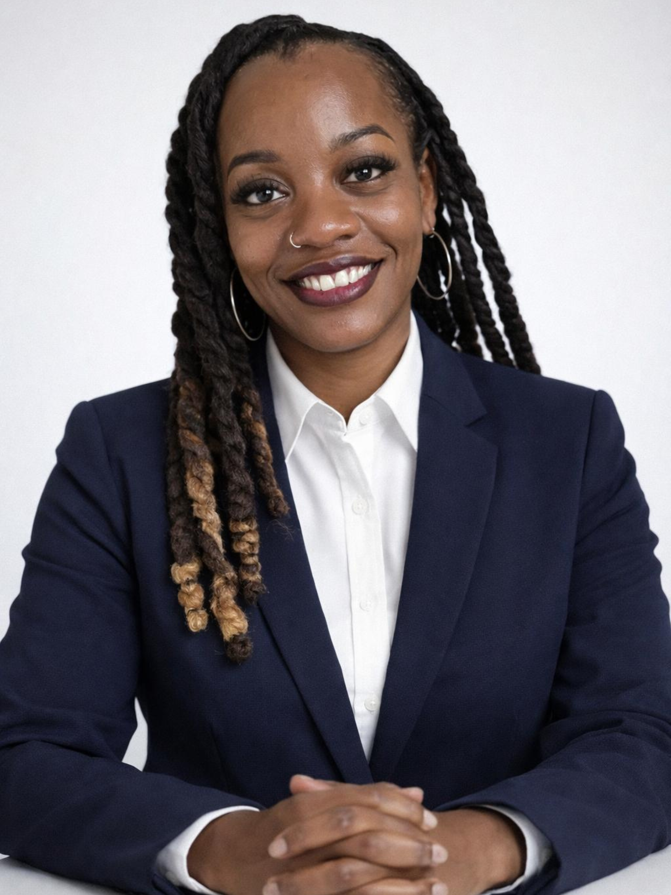 A woman with dark skin and long dreadlocks smiling at the camera, dressed in a navy blazer and white shirt, sitting with her hands clasped in front of her.