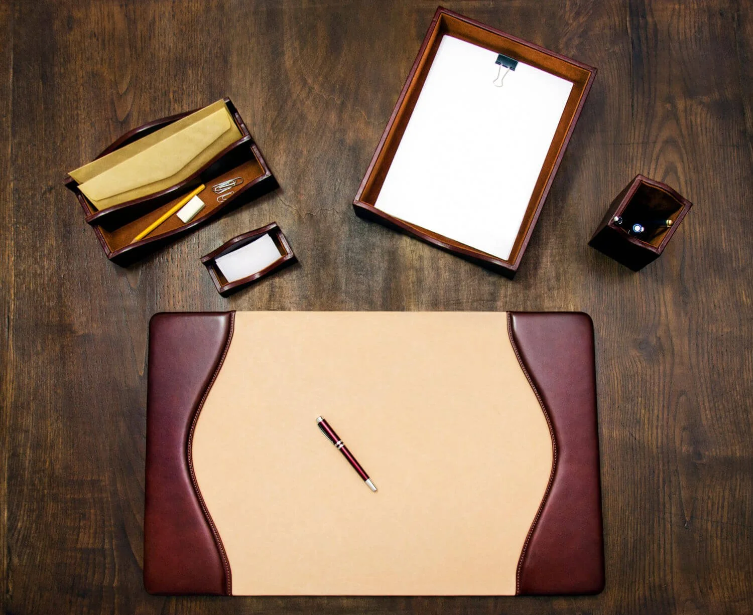 An L-shaped desk pad on a wooden desk with various office supplies including a pen, paperclips, a notepad, and a tray with papers and stationery.