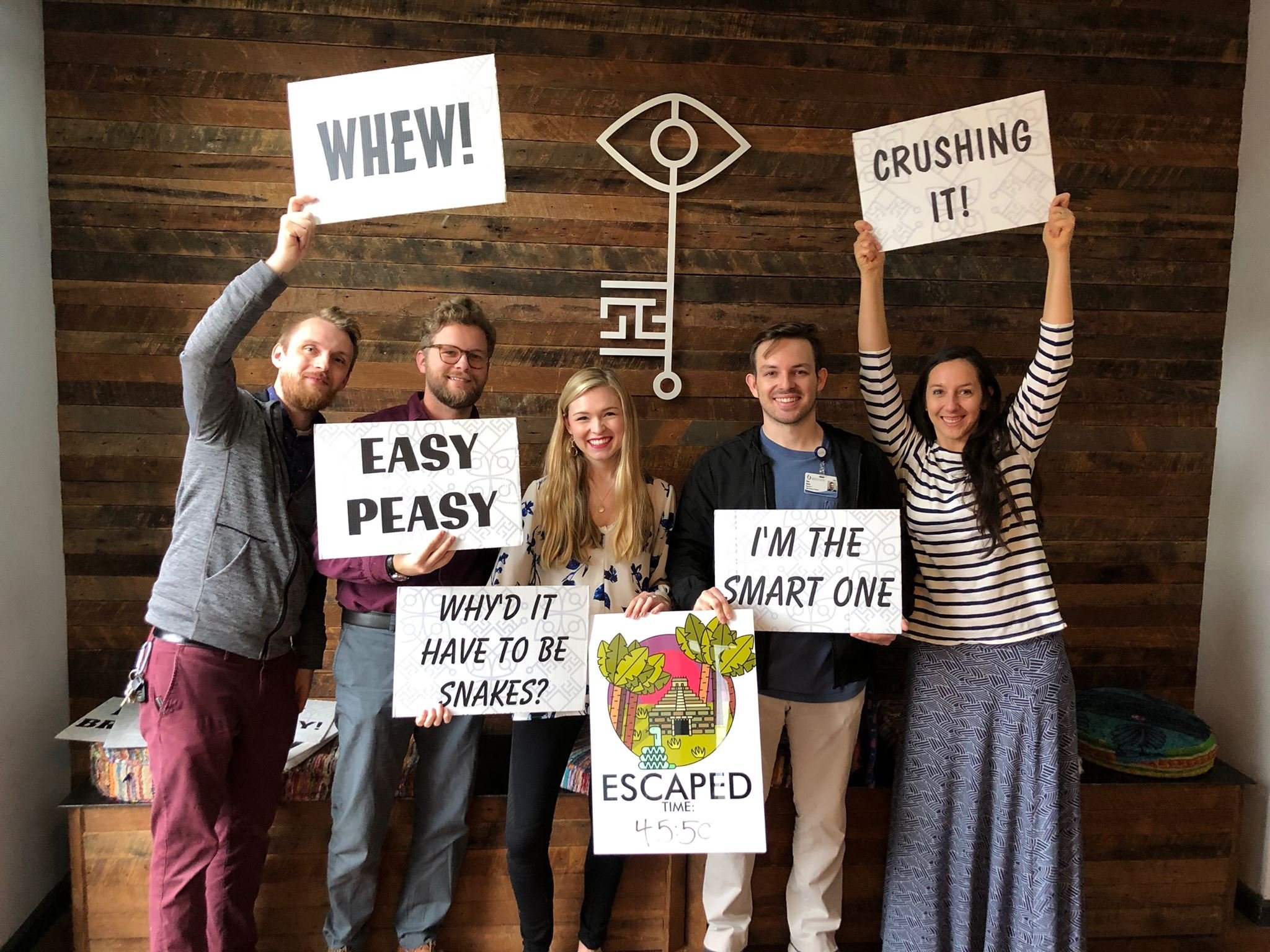 Group of five people holding signs in an escape room, smiling, with wooden wall background and decorative eye symbol