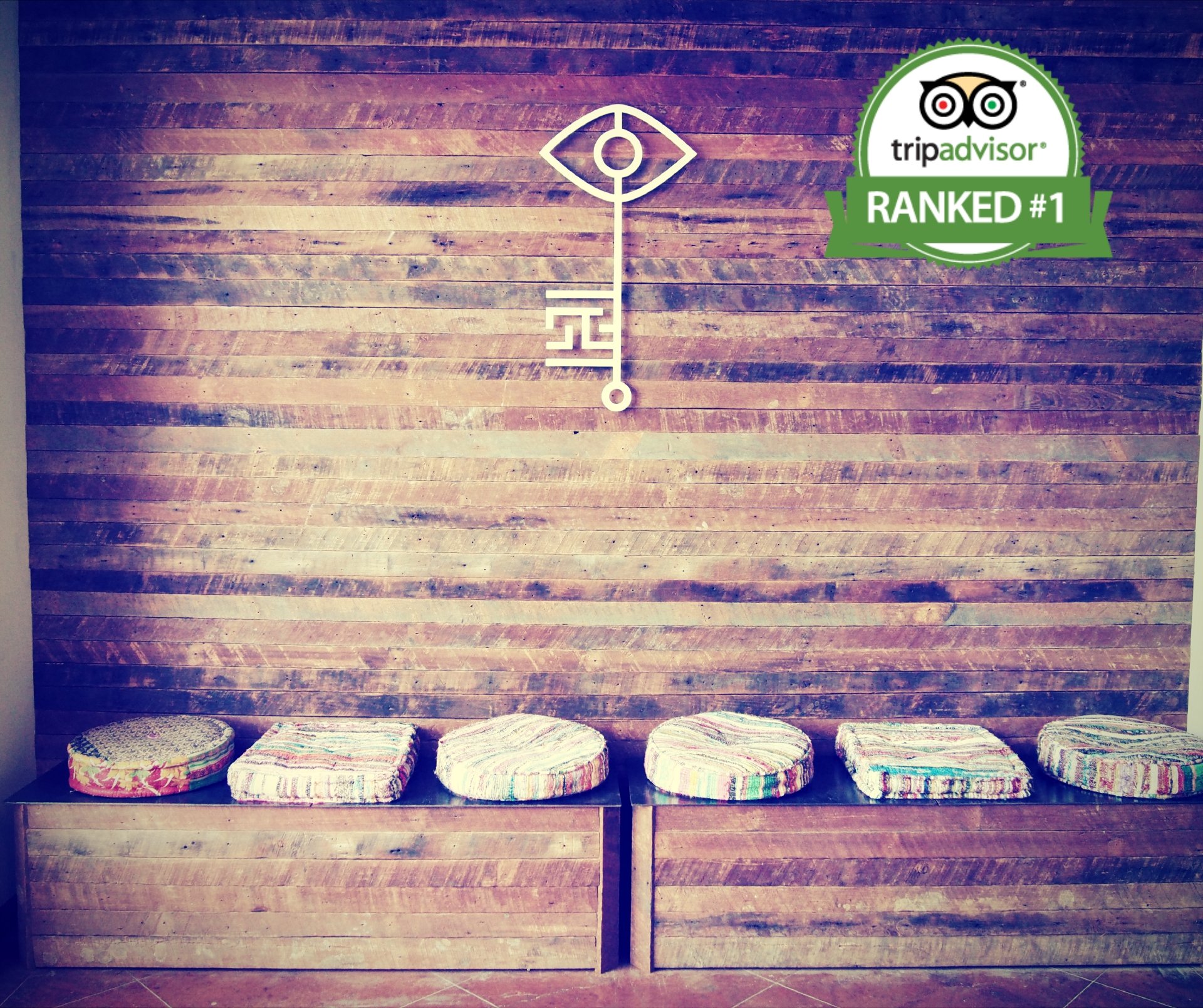 A wooden wall with a minimalist white clock and a TripAdvisor badge indicating a number one ranking. A wooden bench beneath holds six colorful cushions.