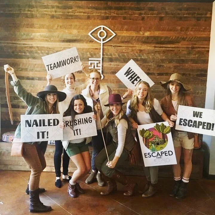 Seven women celebrating their escape from an escape room, holding signs with phrases like 'TEAMWORK!', 'NAILED IT!', 'CRUSHING IT!', 'WHERE?', 'WE ESCAPED!', and a large illustrated exit sign. They are indoors in front of a wooden wall with a decorative key and eye symbol.