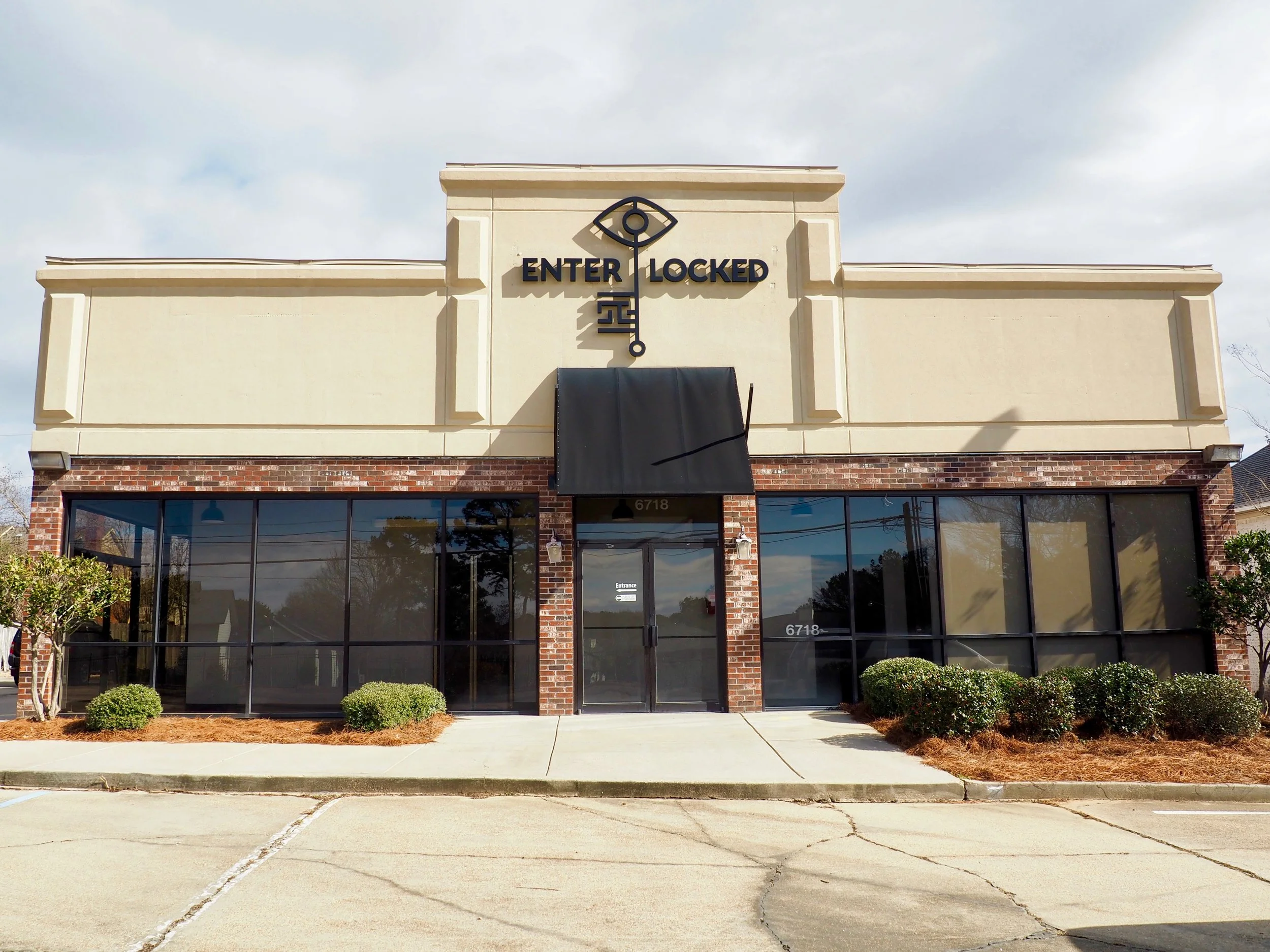 Storefront with a sign reading 'Enter Locked' and a large key symbol on the beige facade, brick accents below, glass windows on either side of a glass door, organized landscaping, and sidewalk in front.