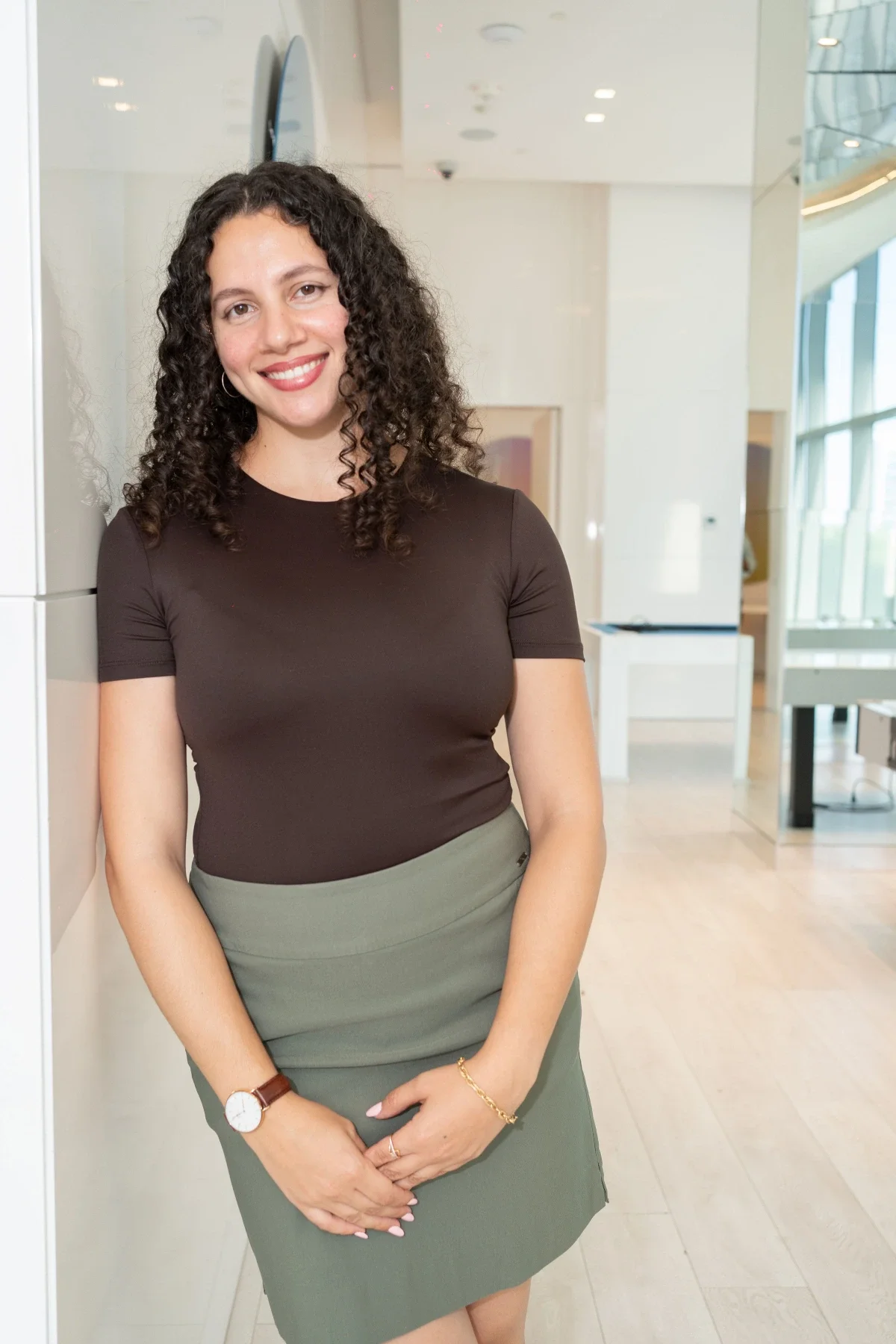 A smiling woman with curly dark hair leaning against a wall in a modern, well-lit building