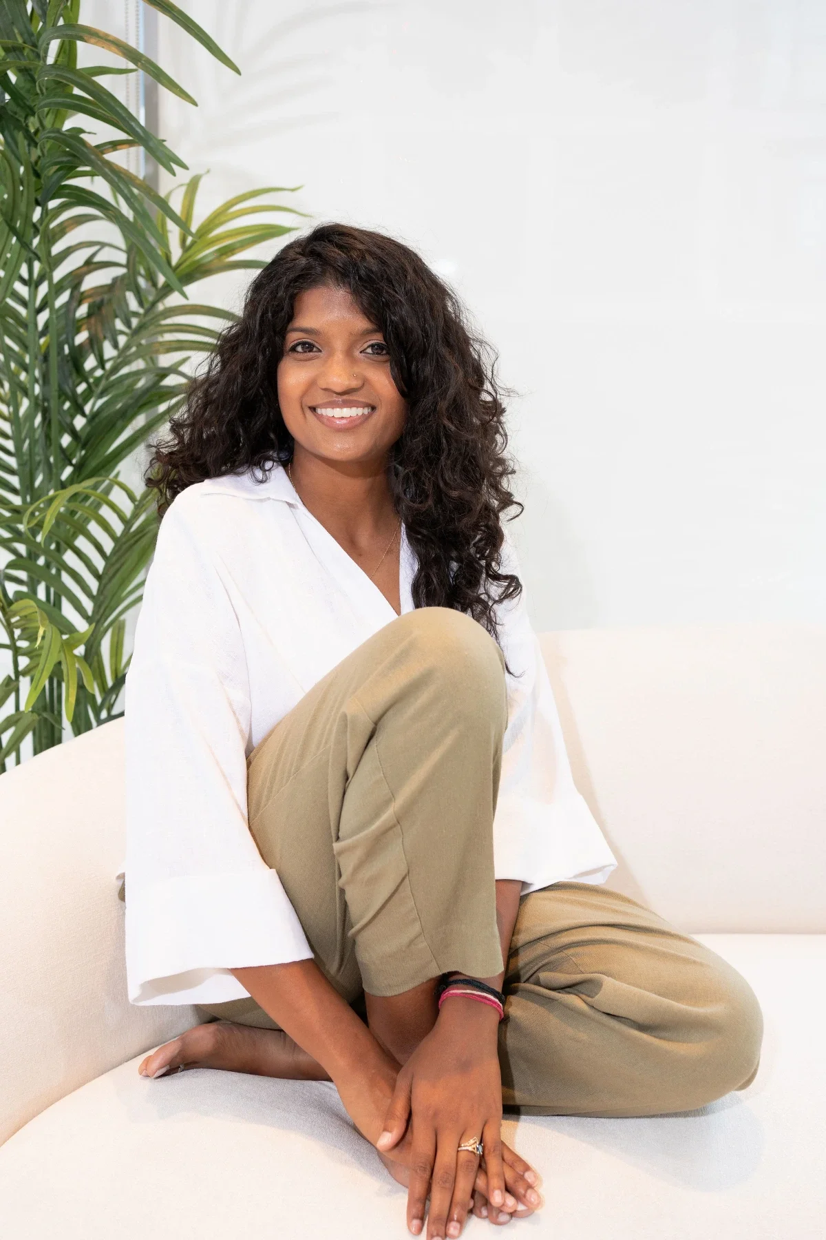 A woman sitting on a white couch with one knee bent and smiling, with a large green plant behind her, in a bright room with white walls.