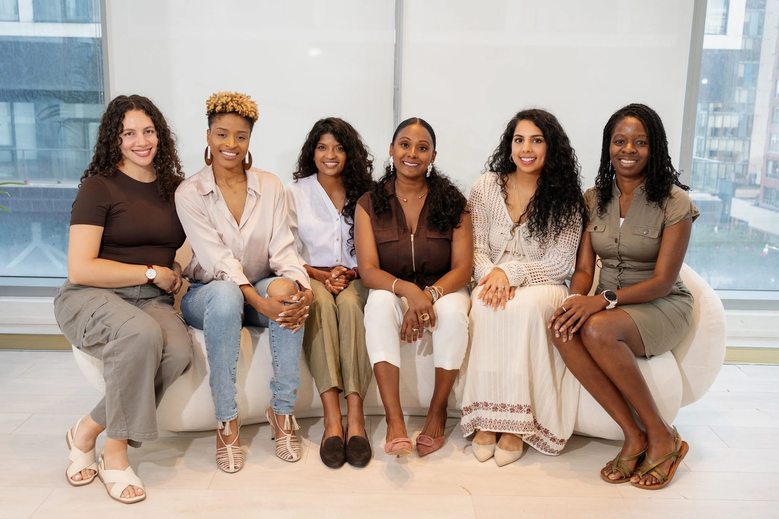 Six women sitting on a white couch in front of large windows in a high-rise building.