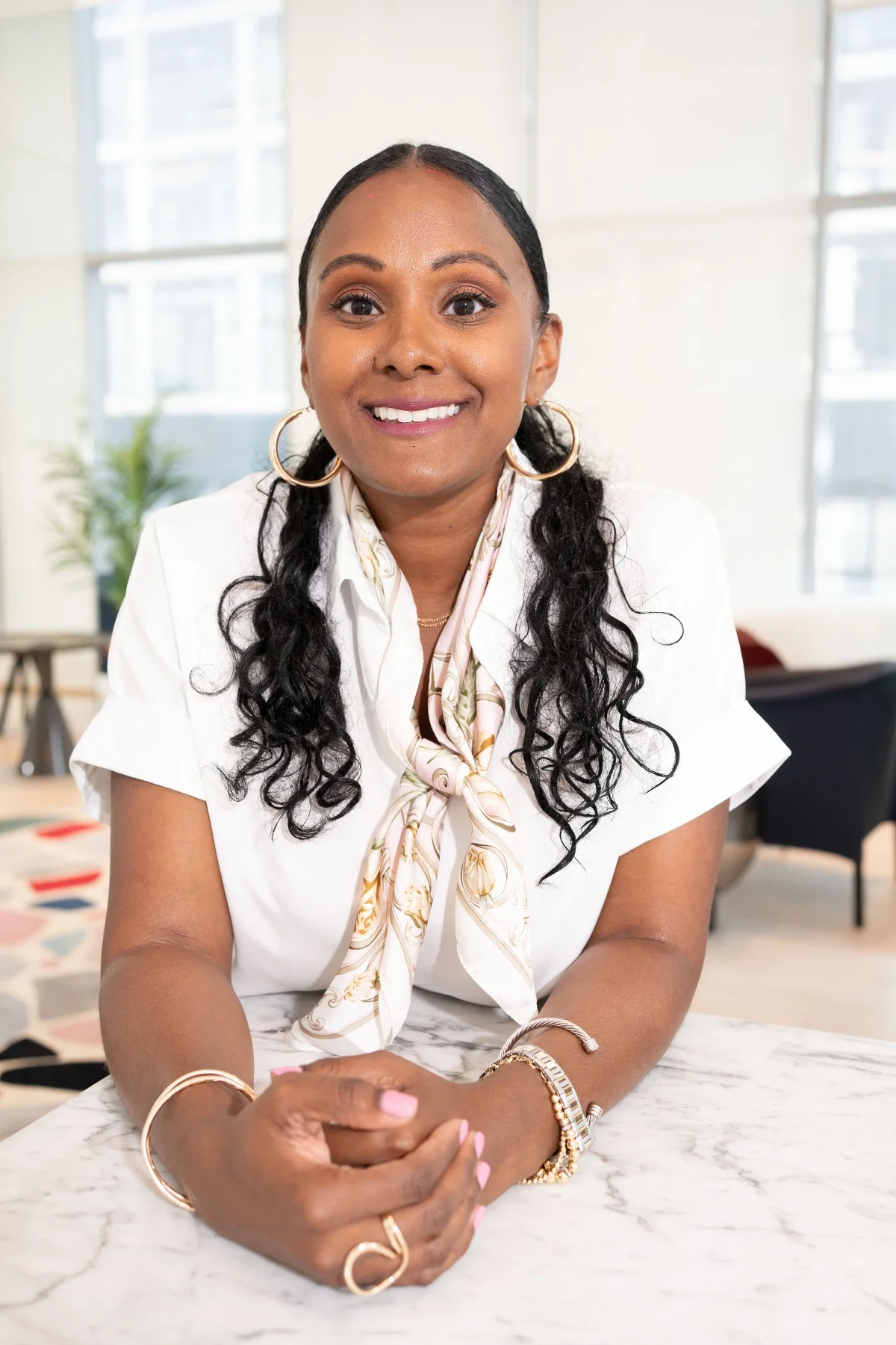 A smiling woman with long, curly black hair, wearing a white blouse, a patterned silk scarf, large hoop earrings, and gold jewelry, is seated at a marble table in a bright, modern office space with large windows and a plant in the background.