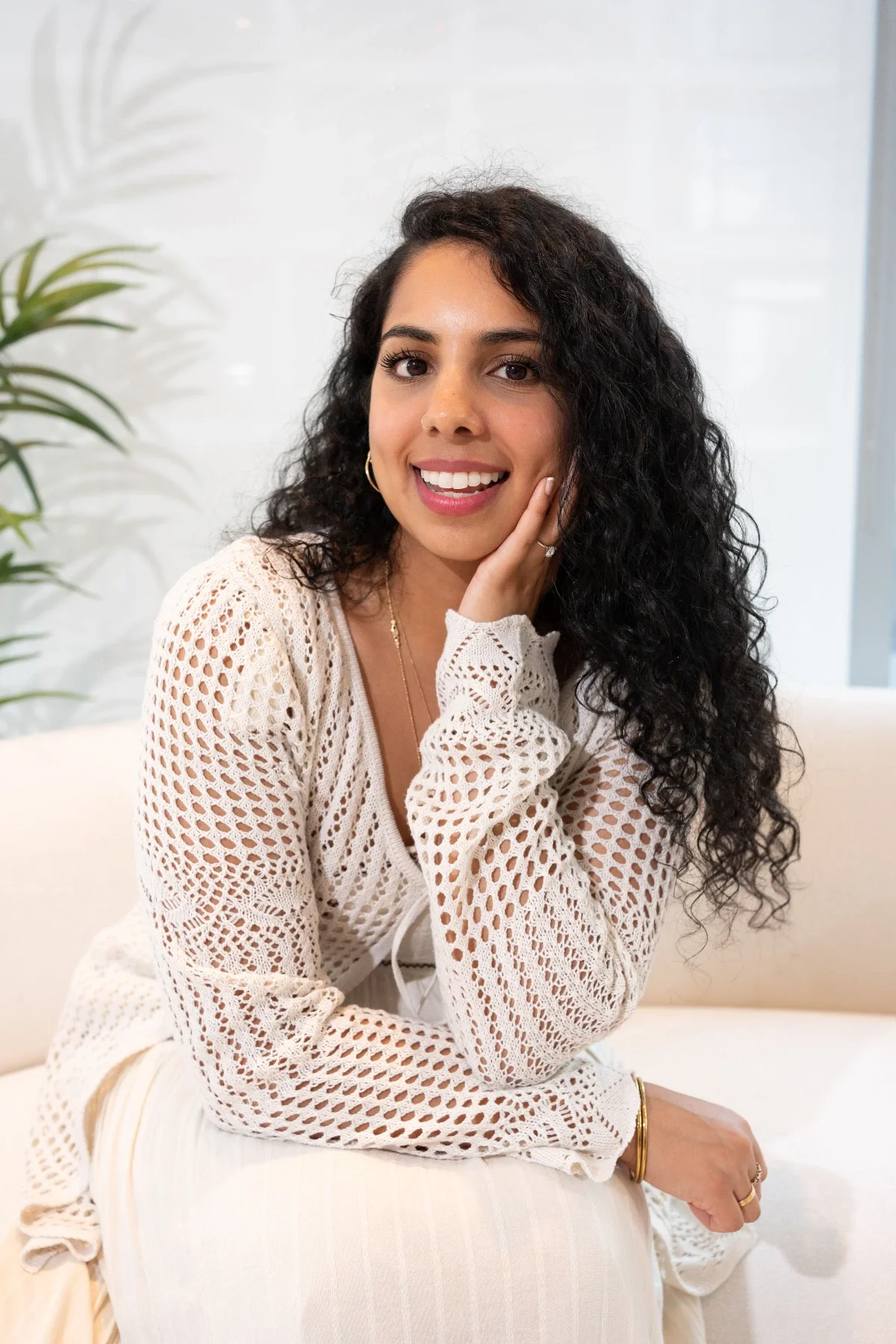 A young woman with curly dark hair, wearing a cream crochet top and gold jewelry, sitting on a white sofa and smiling at the camera indoors.