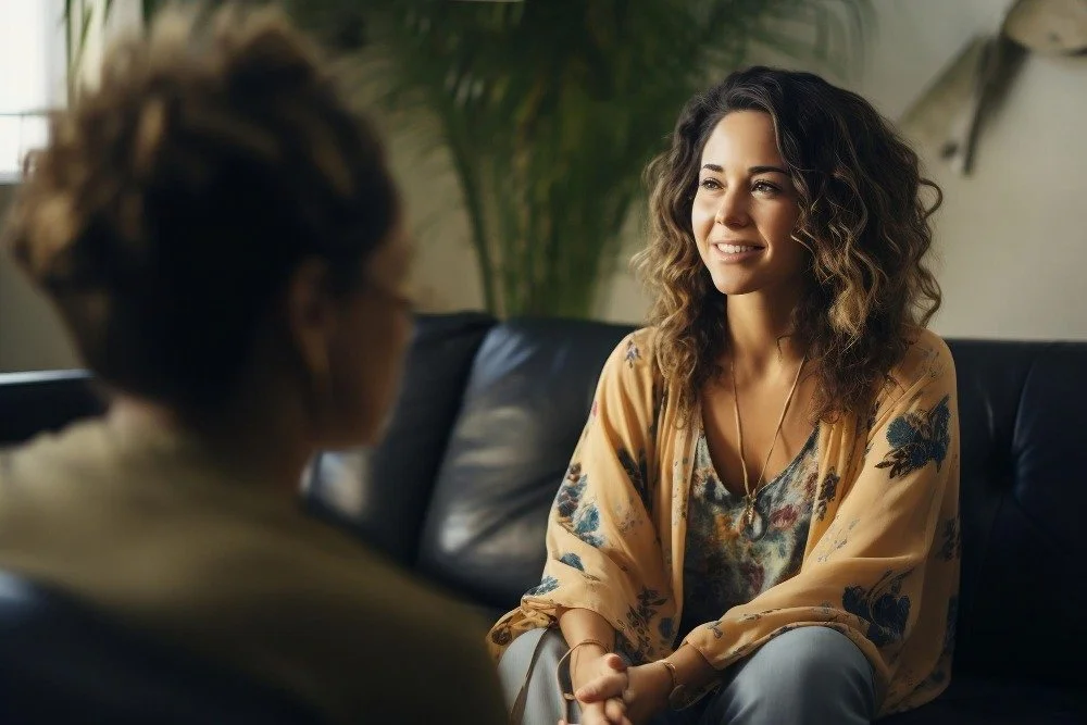 Two women having a conversation in a cozy, well-lit living room with a plant in the background.