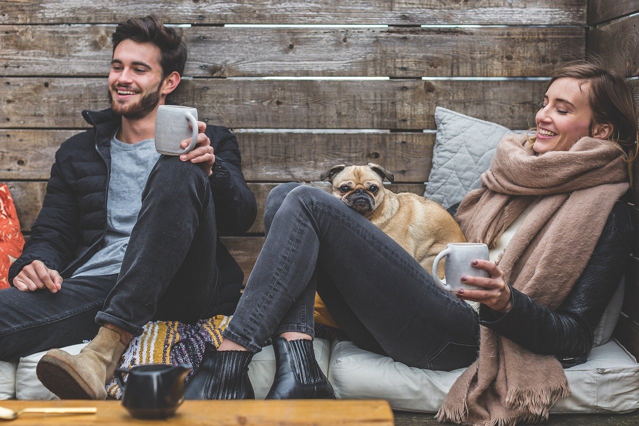 A man and woman sitting on a sofa outdoors, smiling and holding coffee mugs, with a pug dog resting on the woman's lap, in a cozy setting with a wooden wall background.