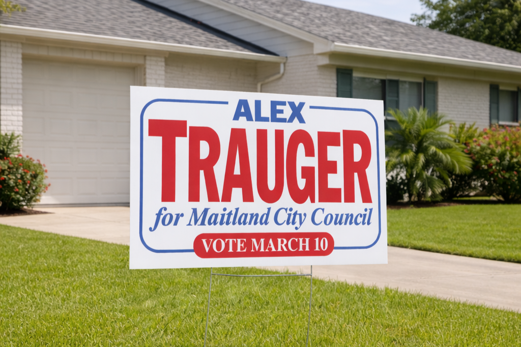 Yard sign promoting Alex Trauger for Maitland City Council, with a house and garden in the background.