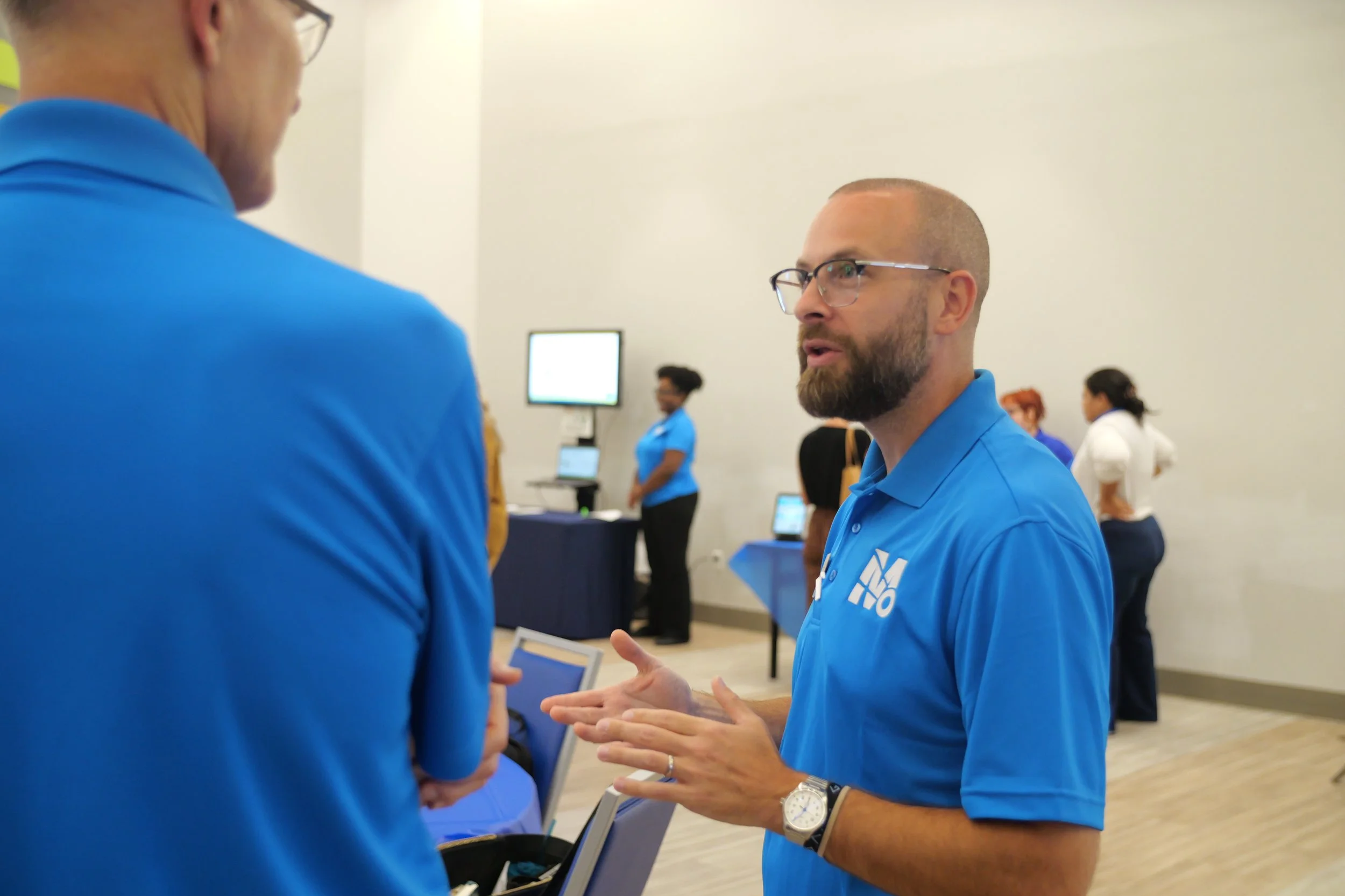 A man with glasses, a beard, and a blue polo shirt with a logo, is speaking and gesturing with his hands in a conversation at an indoor event, with other people and display tables in the background.