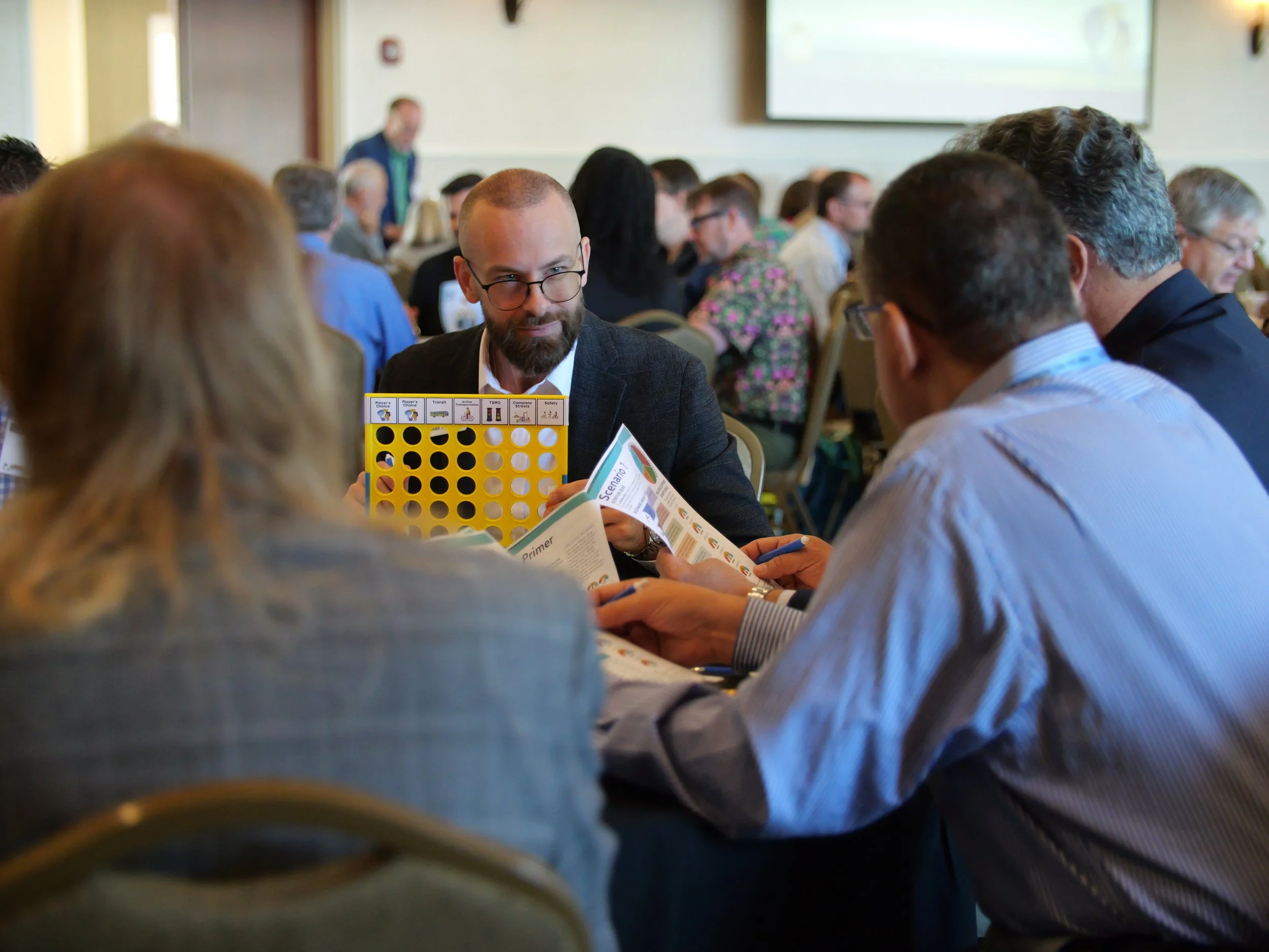Group of people in a conference room engaged in a discussion, with various printed materials and a yellow Connect 4 game on the table.