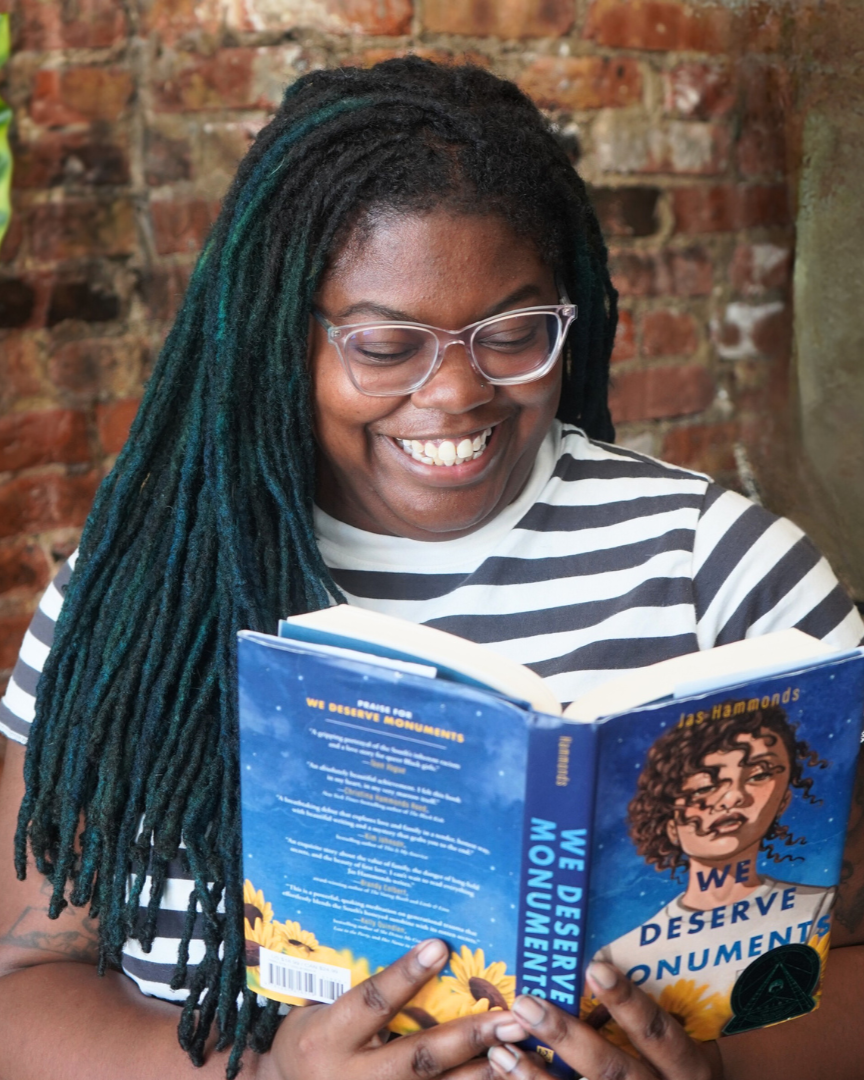 Killian, a young black nonbinary person with long blue locs and clear plastic glasses, reads the book "We Deserve Monuments" by Jas Hammonds. They are smiling at the book contents