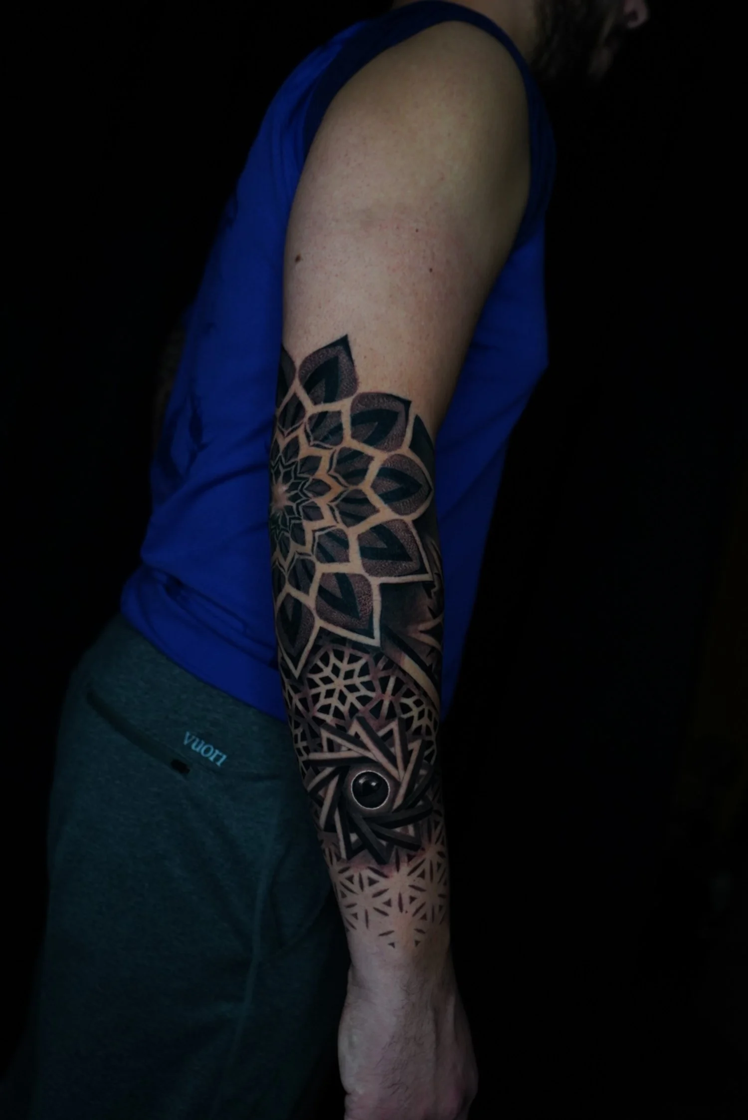 A person showing a detailed geometric tattoo on their forearm, wearing a blue sleeveless shirt and gray pants.