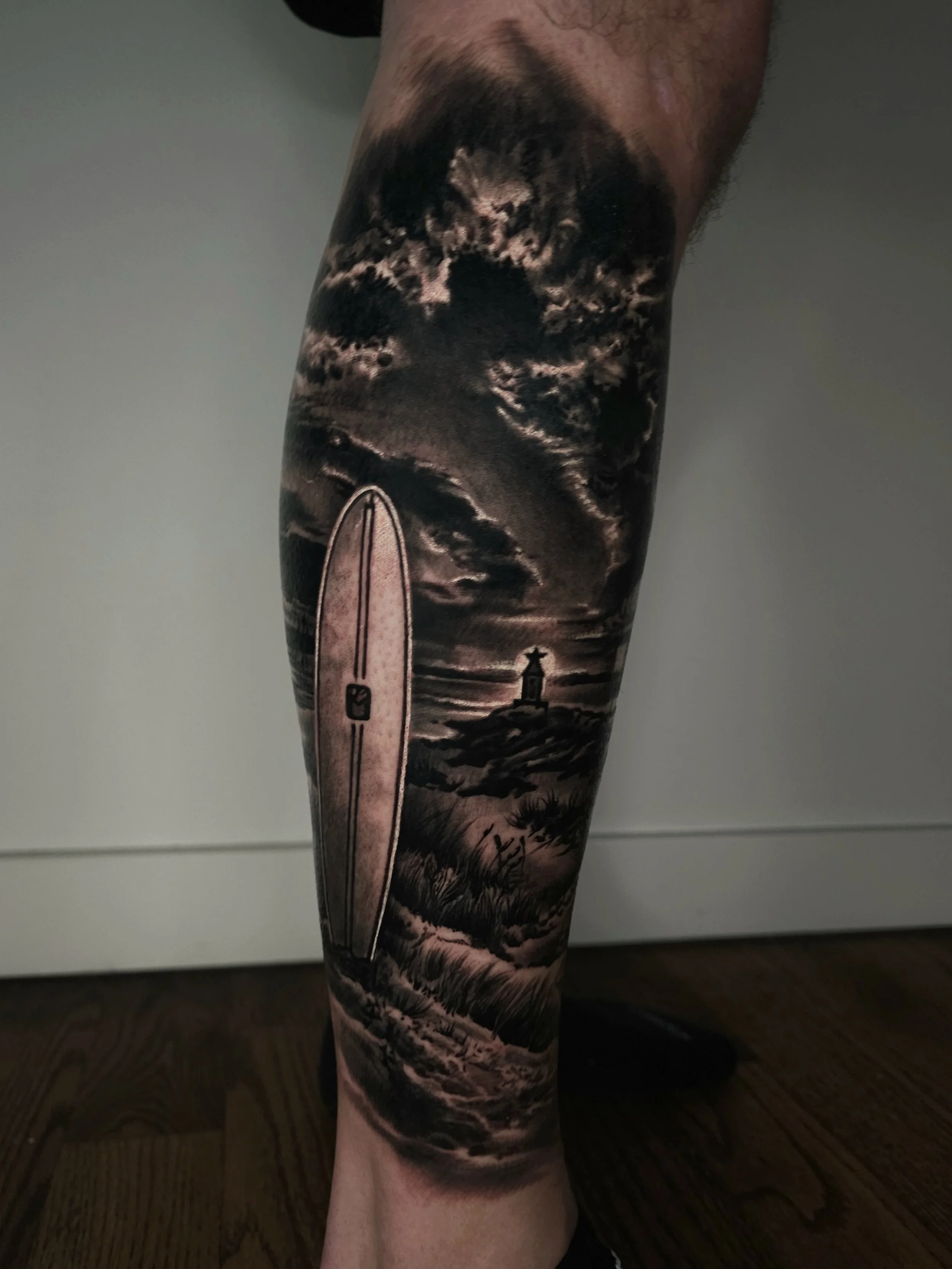 Tattoo of a surfboard with a landscape of cloudy sky and lighthouse on a person's leg.