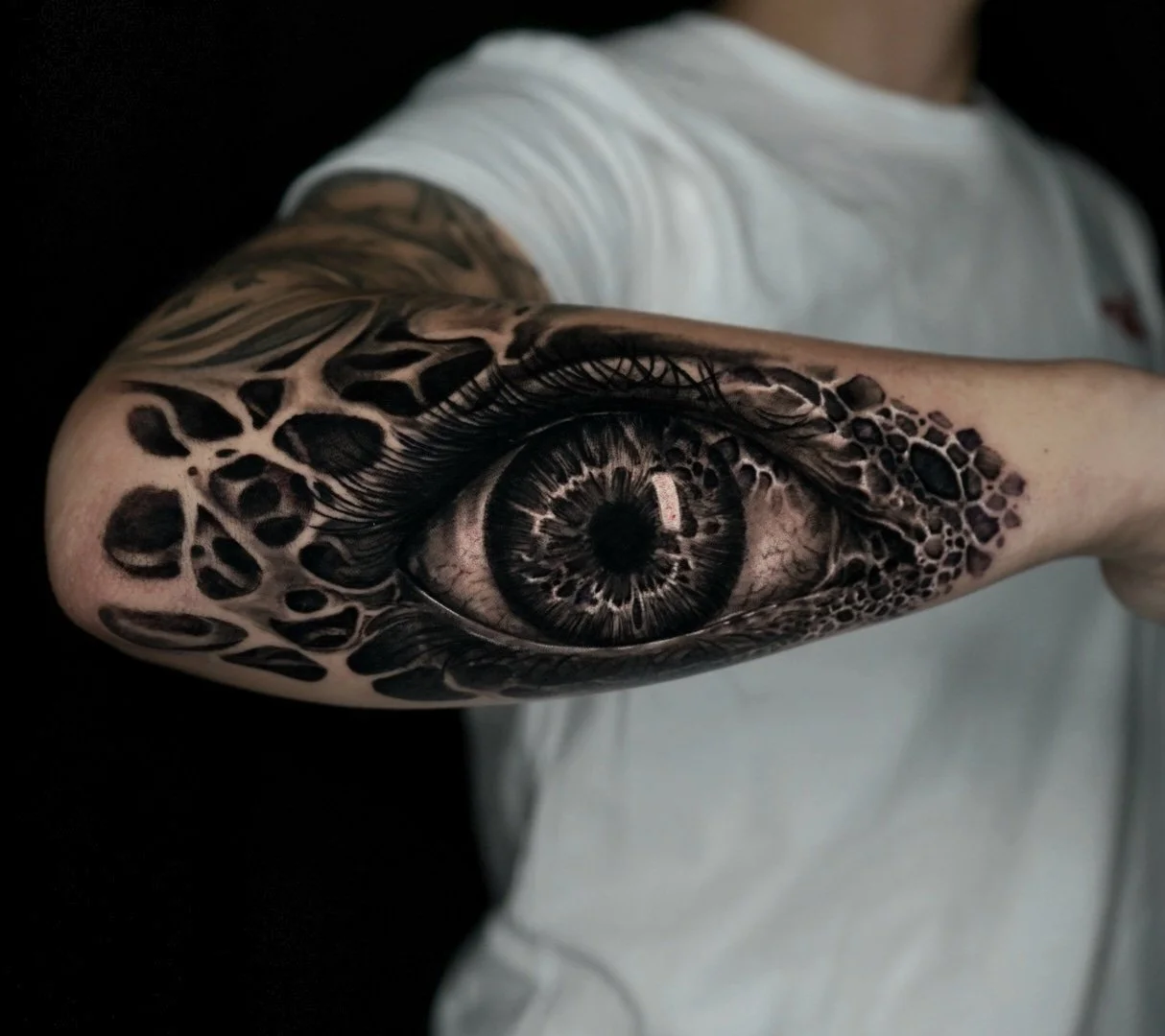 Tattoo of a realistic human eye on a person's arm, with detailed eyelashes and patterns around the eye, against a black background.