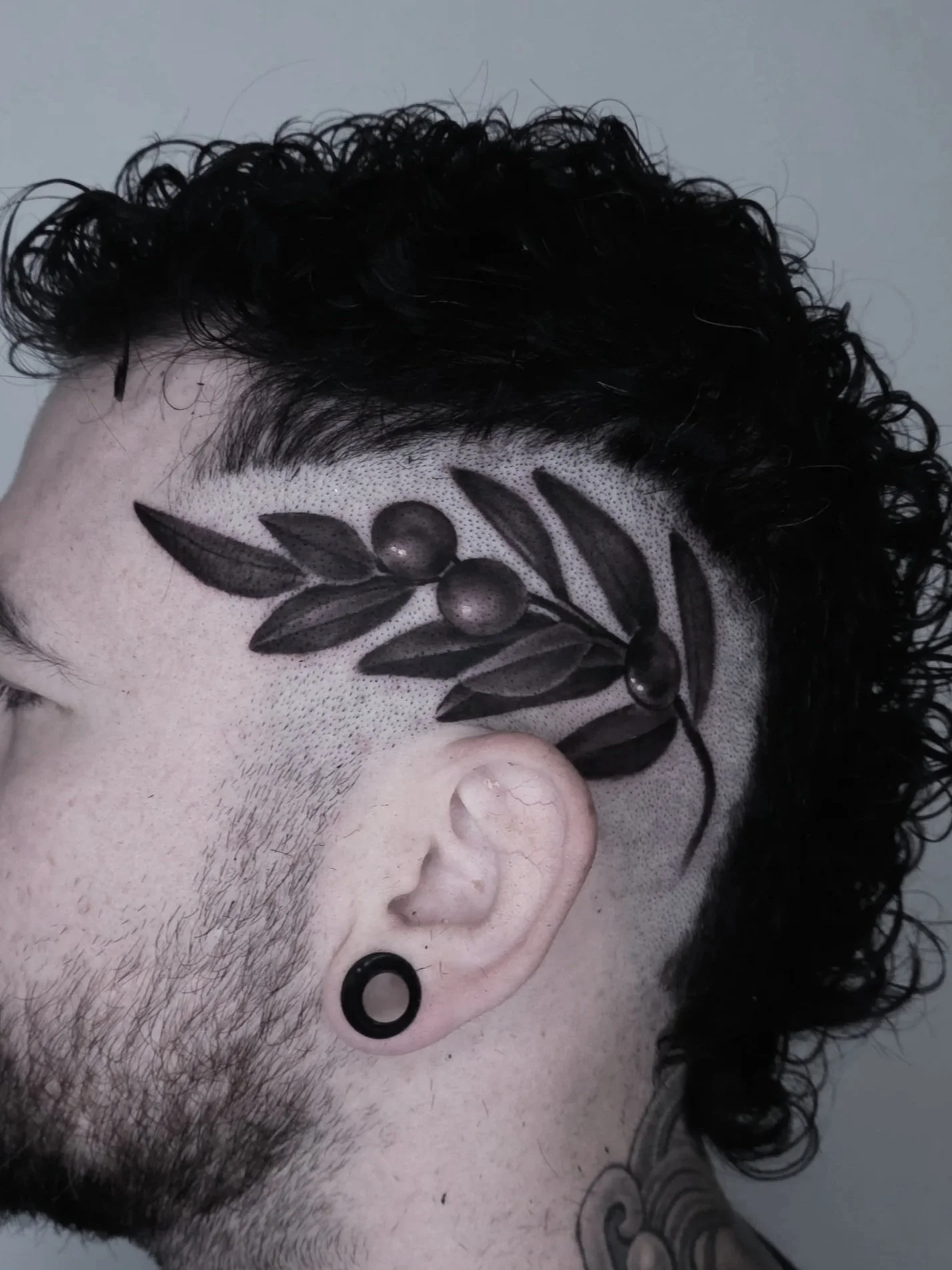 Close-up of a man's head with a unique haircut featuring a black ink tattoo of an olive branch on the side of his scalp. The tattoo includes detailed leaves and three olives, and the man has a black ear gauge earring.