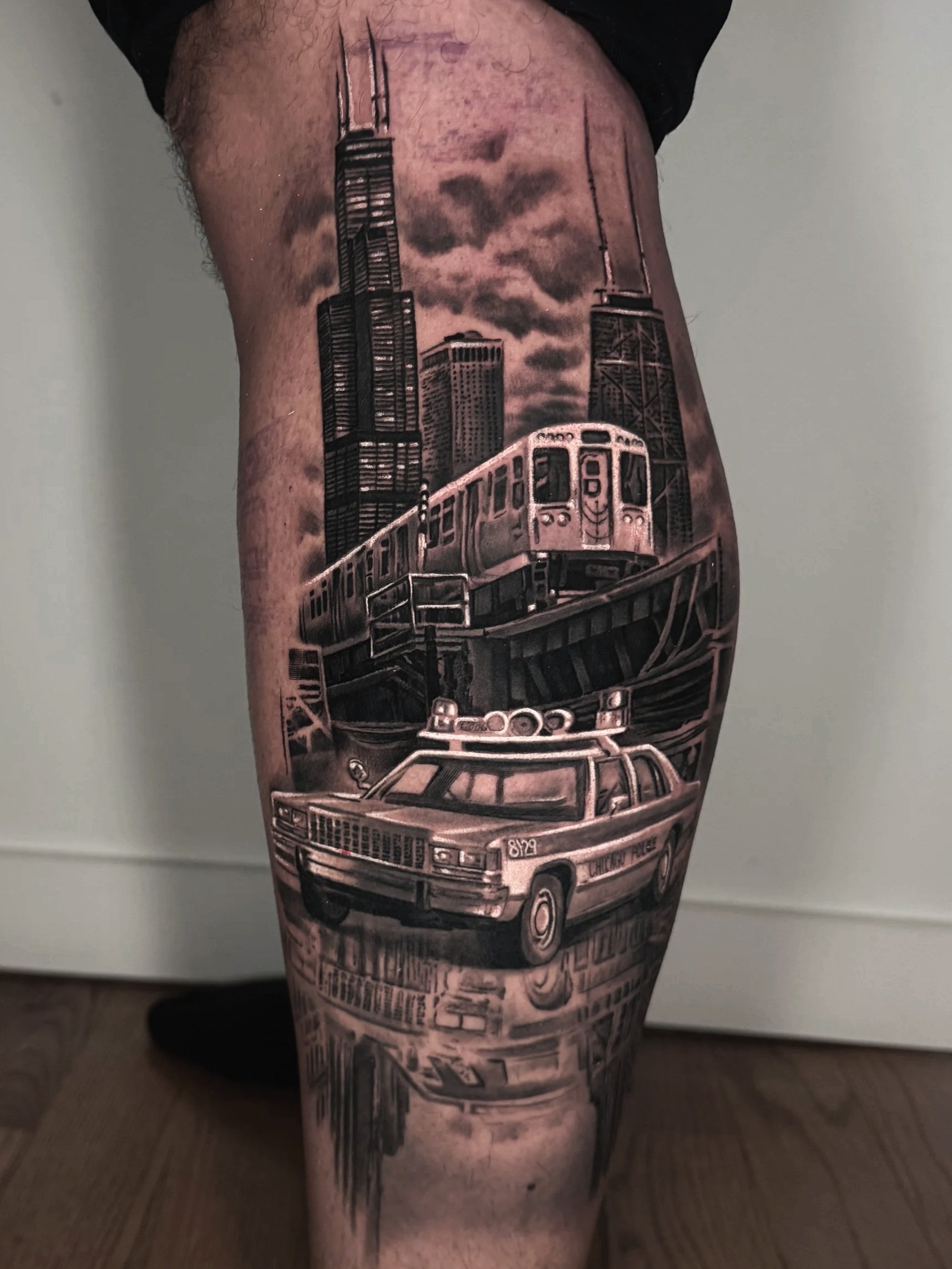 Black and Grey realism tattoo of a Chicago police car, a CTA train, and the Chicago Skyline on outer calf by award winning artist Nelson Aviles with 10 years experience in Chicago at Black Atlas Studios.