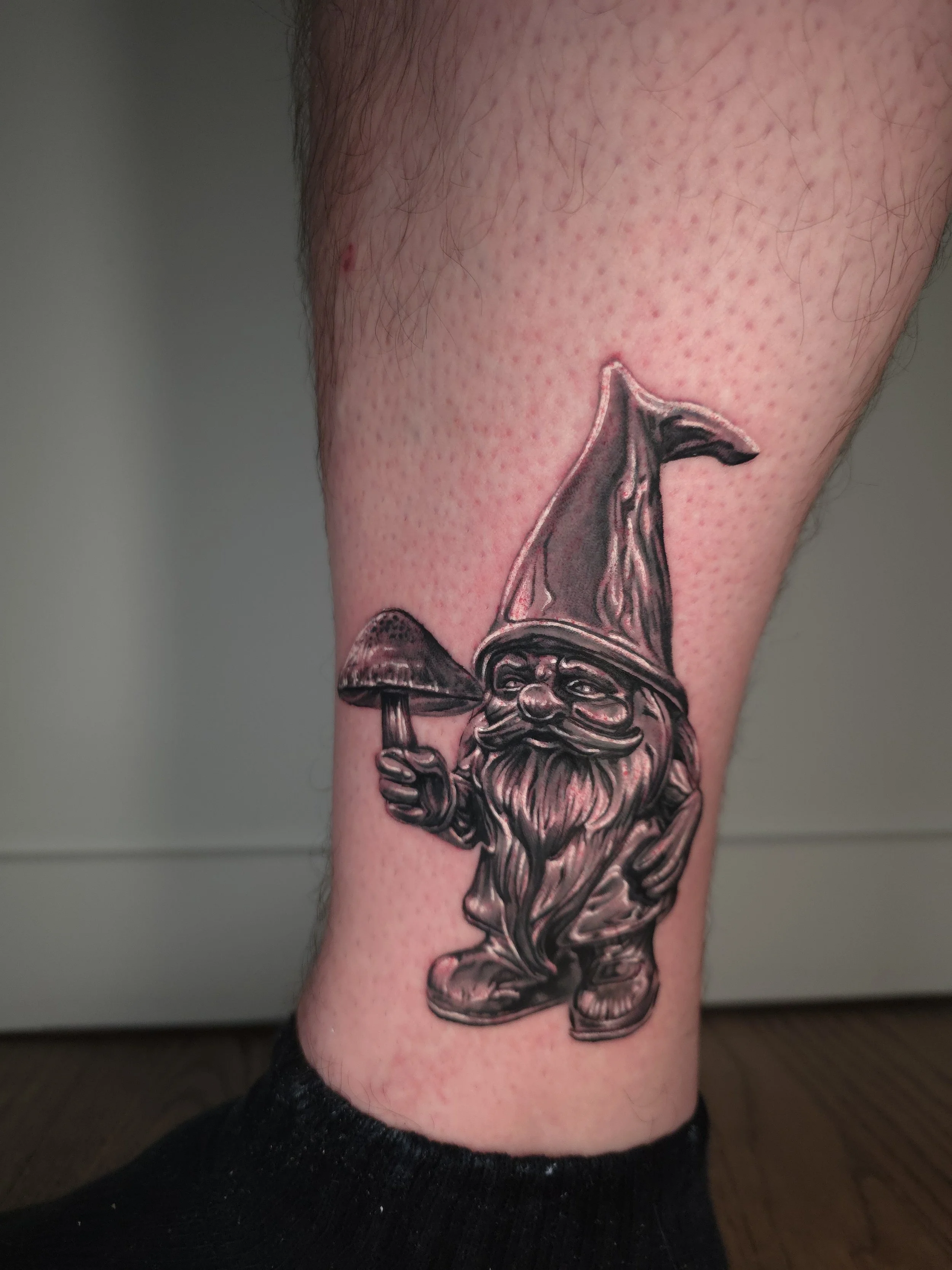Tattoo of a gnome with a beard, wearing a pointed hat, holding a mushroom, on a person's calf.