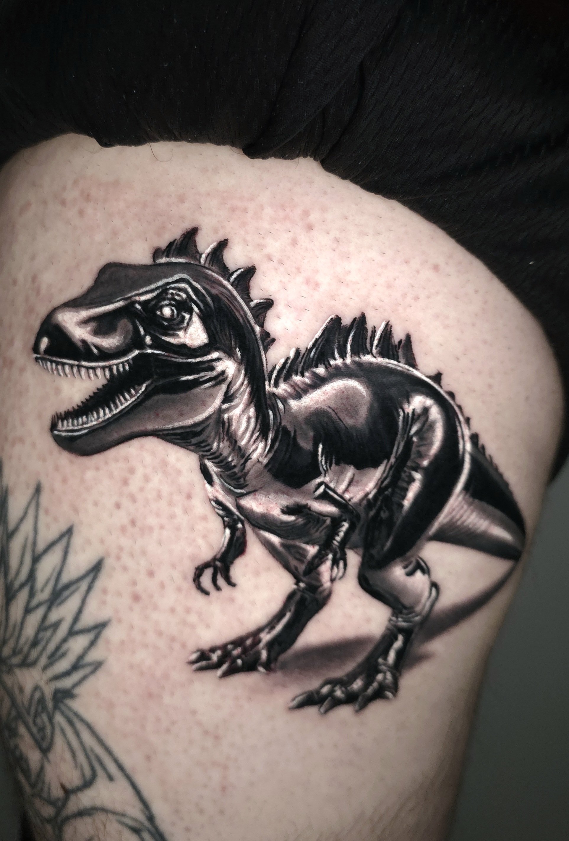 Black and gray tattoo of a dinosaur on a person's skin, with a detailed head, open mouth, and sharp teeth.