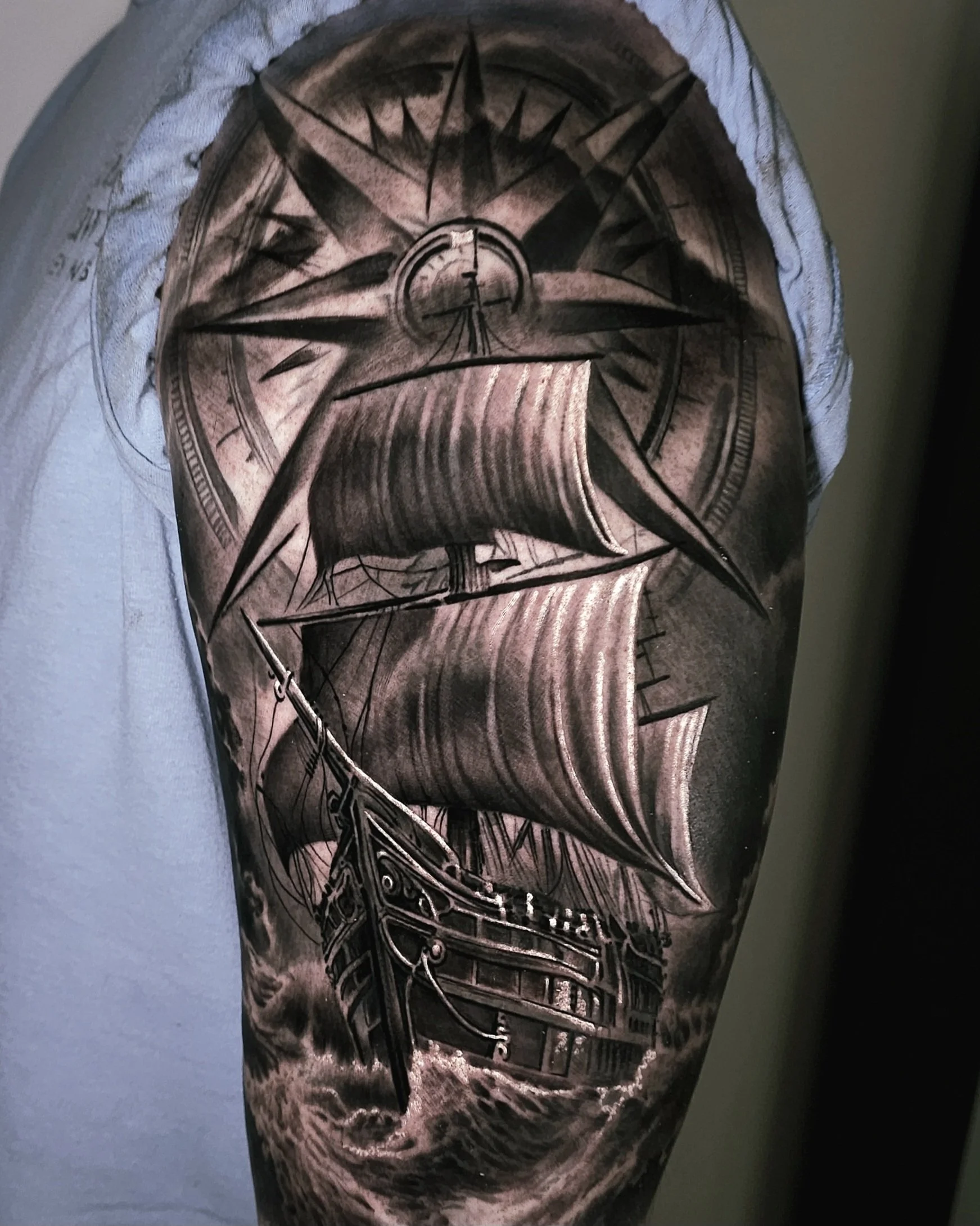 Black and gray tattoo of a sailing ship with large sails, a compass rose at the top, and stormy waves below.