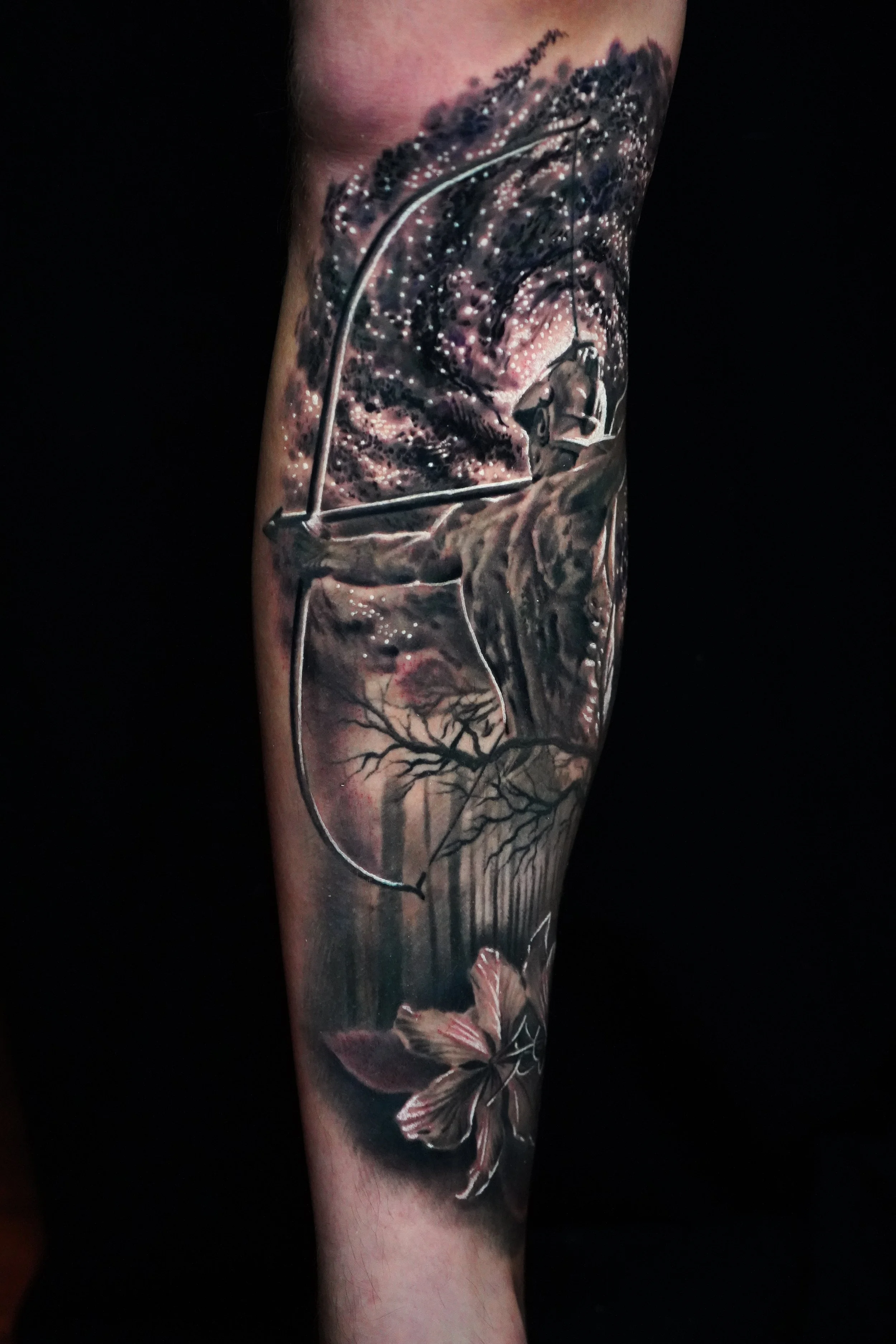 Detailed black and gray tattoo featuring a bow and arrow, a flipped butterfly, a praying mantis on a branch, trees, and a starry sky with a galaxy, covering a person's forearm.