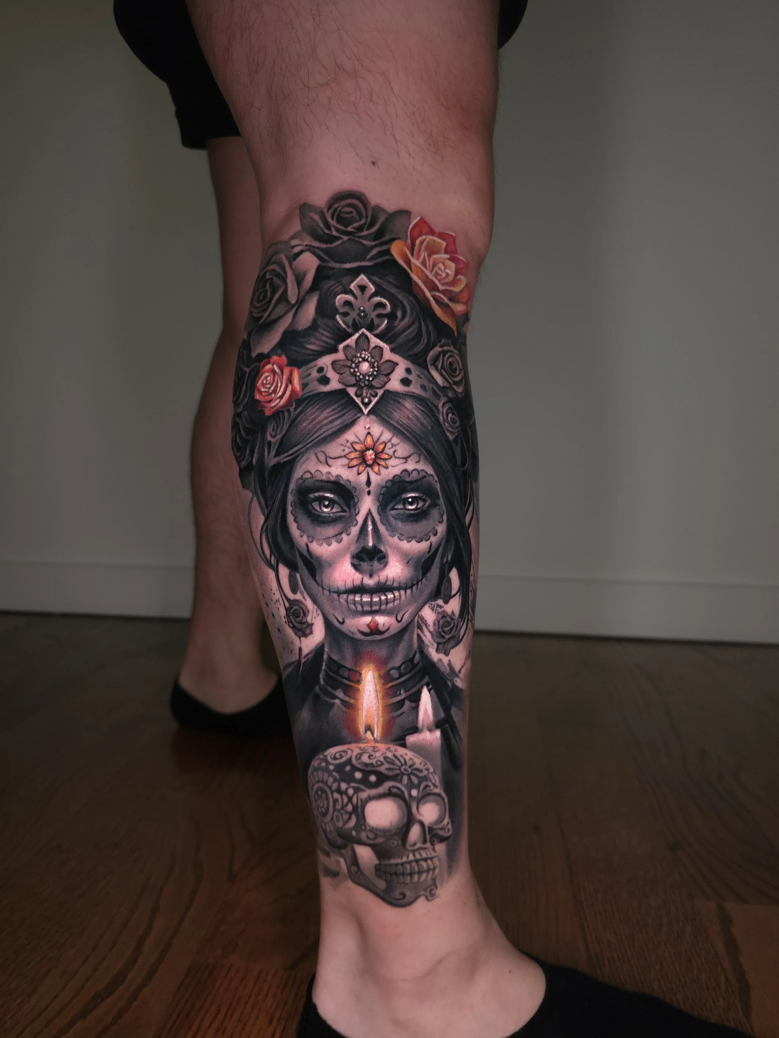 Colorful tattoo on person's lower leg featuring a woman with a sugar skull face, surrounded by roses, candles, and decorative elements.