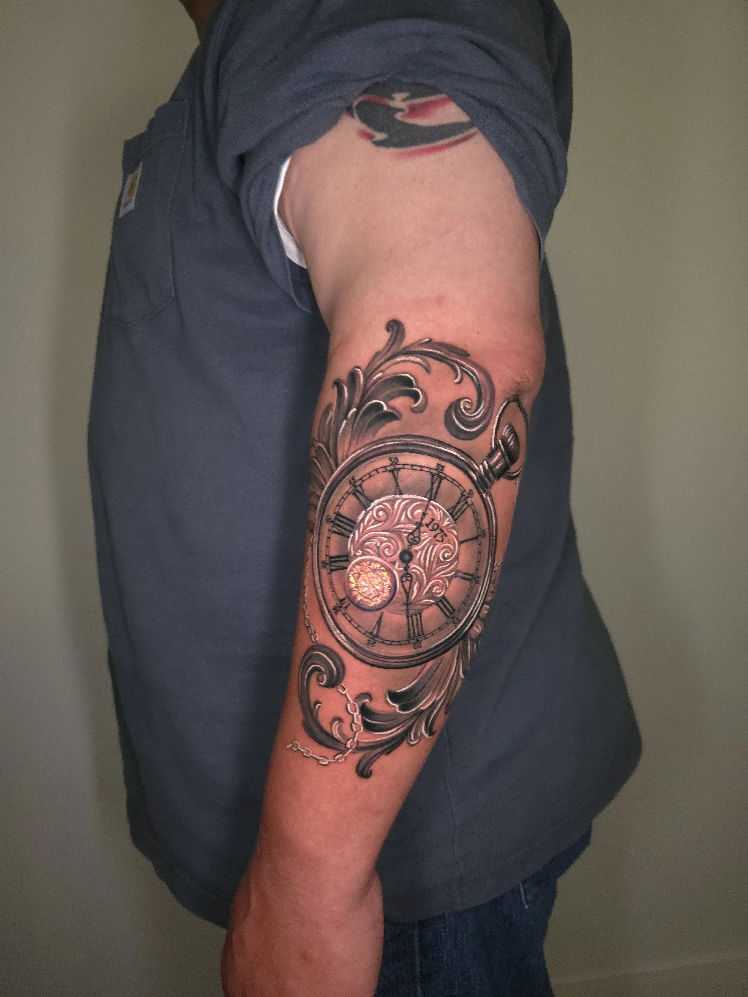 Tattoo of a vintage pocket watch with Roman numerals and intricate design, surrounded by decorative swirls and chains, on a person's arm.